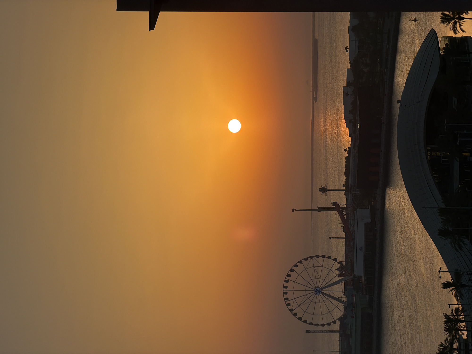 An overhead, vertical, medium shot captures a vibrant sunset over a city waterfront. The sun, a bright, white orb, sits low in the orange sky, casting a shimmering reflection on the water. In the mid-ground, a large Ferris wheel stands silhouetted against the luminous sky, to its right, there is a long cargo ship on the water and more buildings are visible along the coastline. In the foreground, a dark, angular building juts into the frame from the right side. Palm trees line the waterfront.