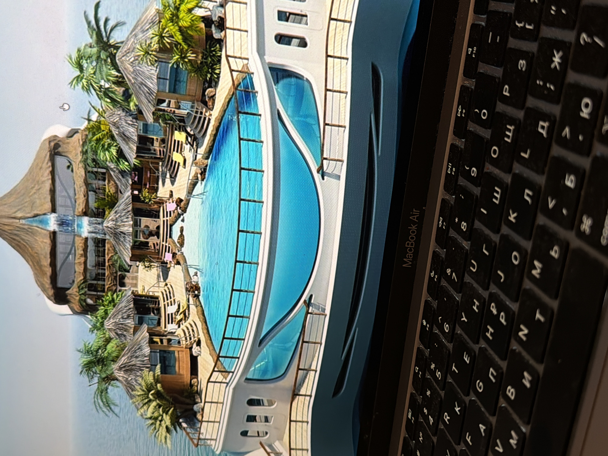 The image captures a close-up, angled view of an open MacBook Air laptop, with its screen displaying a vibrant digital rendering of a luxurious tropical resort or island. The screen content features several thatched-roof bungalows or villas nestled among lush green palm trees, overlooking a serene body of blue water, likely the ocean. A prominent feature is a modern, white floating structure, possibly a large yacht or a specialized resort section, with an elongated, curved swimming pool integrated into its design. A small waterfall can be seen near some of the huts. A mouse cursor, shaped like a pointing hand, is visible on the screen.
The right side of the image shows a portion of the laptop's black keyboard, which appears somewhat dusty or worn. The keys are imprinted with both Latin and Cyrillic characters, suggesting a non-English speaking region. Text visible on the laptop includes "MacBook Air" on the bezel. Various alphanumeric and symbol keys are discernable on the keyboard, including function keys F7, F8, F9, F10, and Cyrillic letters like 'Ю', 'Б', 'Ь', 'Т', 'И', 'М', 'П', 'Р', 'О', 'Л', 'Д', 'Ж', 'Э', 'Я', 'Ч', 'С'.
The image is captured from an overhead perspective, with the laptop screen oriented vertically, creating an immersive view of the tropical scene that contrasts with the tangible presence of the device. The digital render depicts a bright, sunny day, enhancing the idyllic atmosphere of the portrayed location.