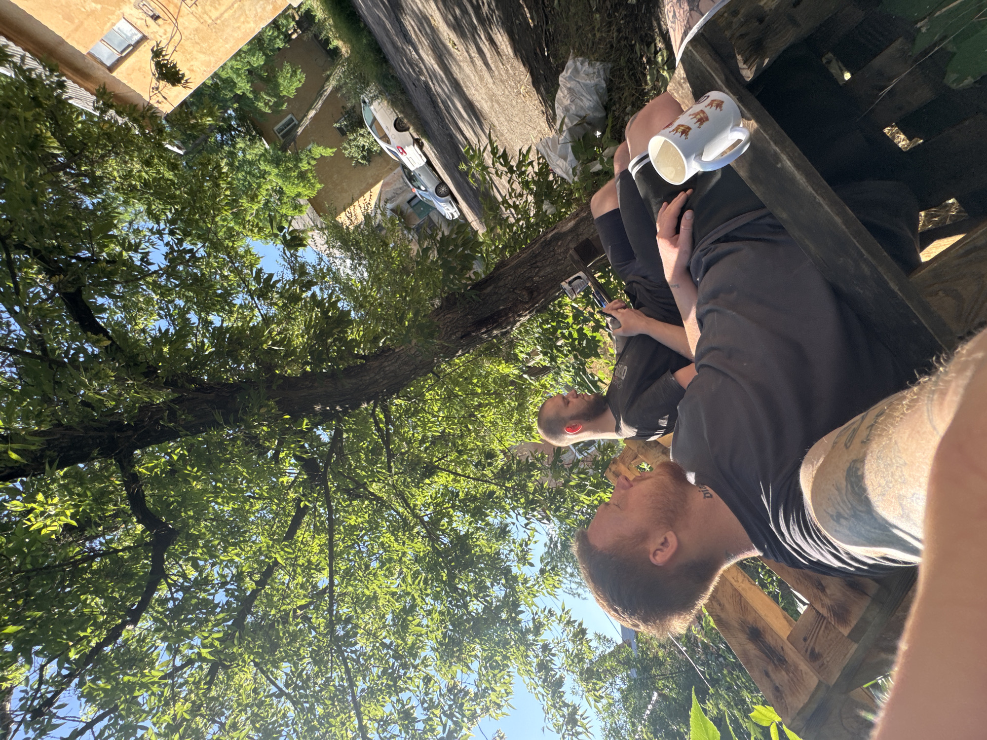 Two men are relaxing outdoors on a sunny day. The man in the foreground, with reddish-brown hair and a beard, is wearing a dark t-shirt and has tattoos on his arm. He is looking upwards and to the right. He has a tattoo on his neck. The man in the background, with a shaved head, is wearing a dark t-shirt and shorts. He is looking down at his phone. They are seated on a wooden structure that appears to be made of pallets. A white mug with a crown design is placed on the wooden structure near the man in the foreground. The background shows a tree with green leaves, a road, two parked cars, and buildings. The sky is blue.