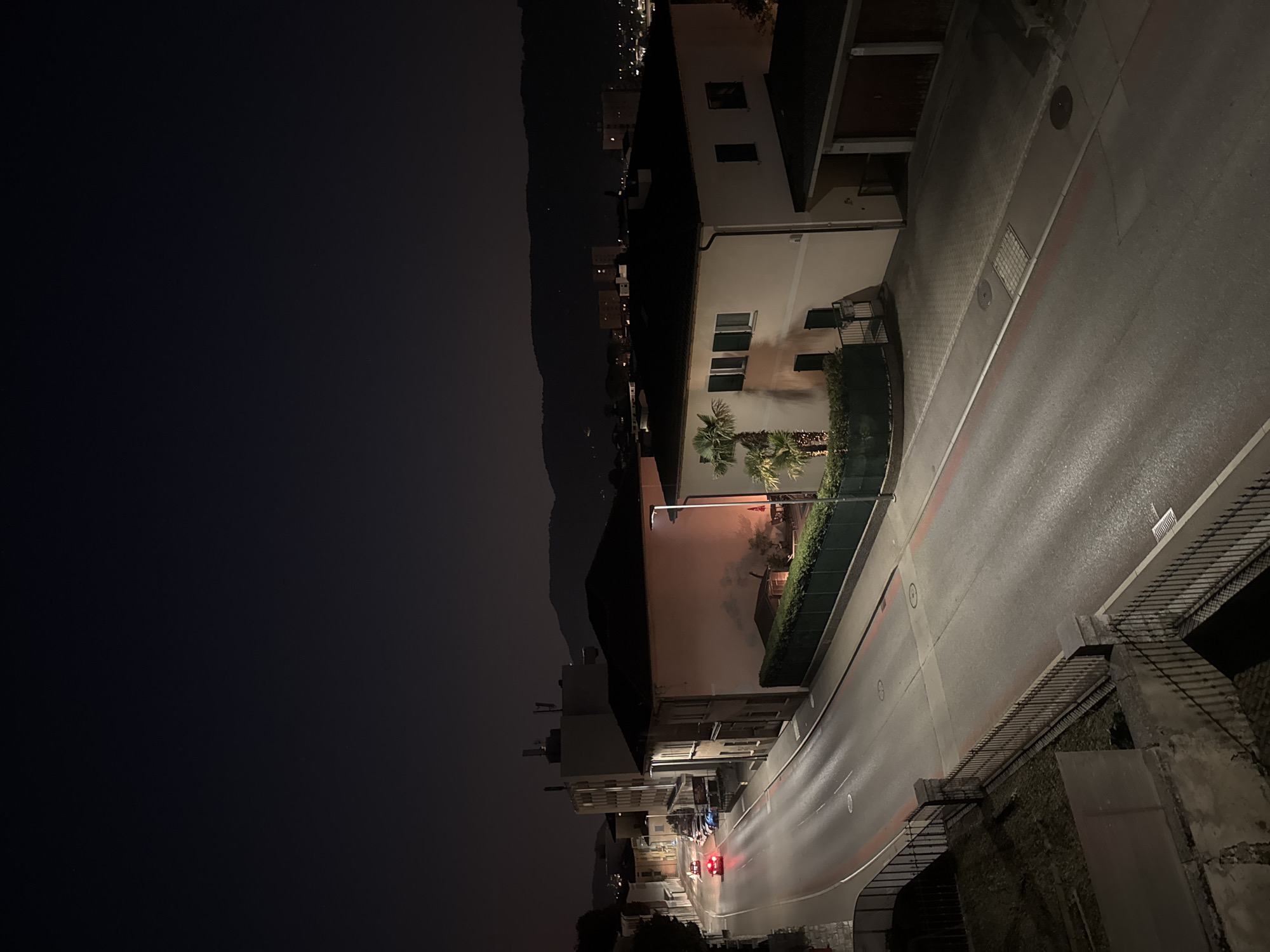 The image captures a nighttime street scene in Balerna, Switzerland. A gently curving road, illuminated by streetlights, dominates the foreground and midground. The road surface appears wet, reflecting the artificial light. Red lines mark the edges of traffic lanes, and several circular manhole covers are visible. A dark metal fence runs along the left side of the road, separating it from a slightly raised, dark grassy area.
On the right side of the road, a light-colored building with dark-shuttered windows stands prominently. A modern streetlight illuminates the area in front of this building. A well-maintained hedge borders the property. To the right of this building, a structure with a dark roof and what appear to be garage doors is visible. Further down the street, several vehicles are parked.
In the background, other buildings and streetlights indicate a residential or commercial area. Beyond the built environment, the silhouette of a dark, tree-covered hill or mountain range is visible against the night sky. The sky itself is dark, with a faint gradient of light towards the horizon, suggesting a clear night. Two cars are visible on the road, one with its red taillights illuminated, heading away from the viewer. One car is red, and appears to be moving away from the viewer, with its taillights illuminated.