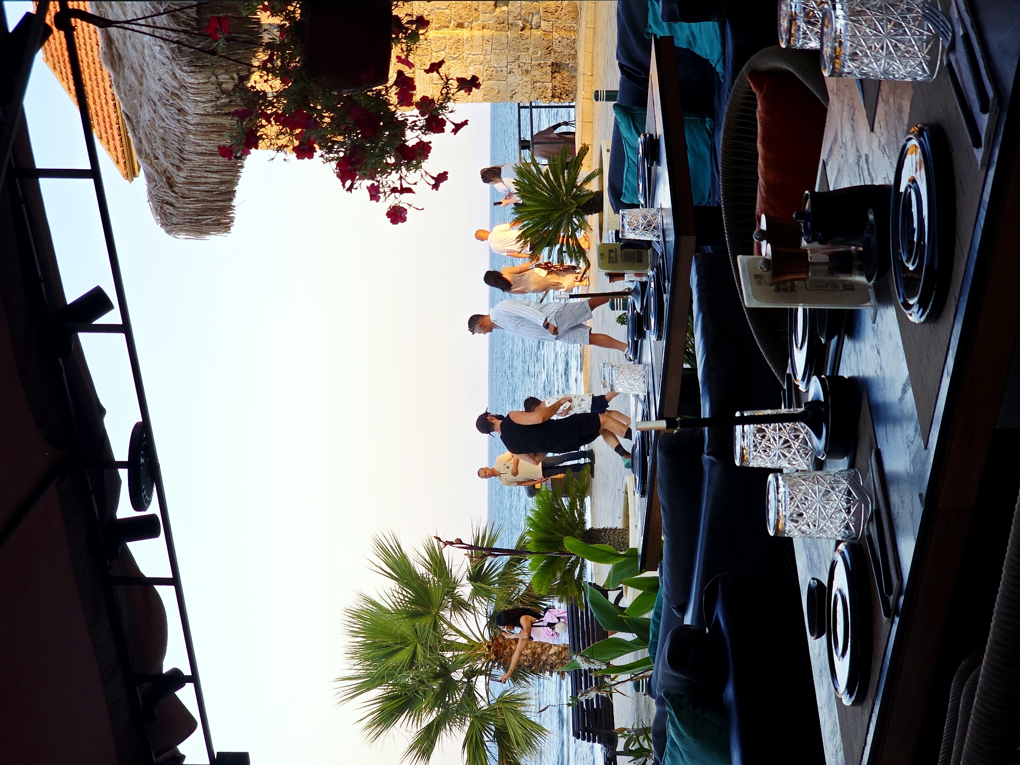 The image depicts an outdoor seating area of a restaurant overlooking the Mediterranean Sea, likely in Larnaca, Cyprus. In the foreground, tables are set with black plates, cutlery, and ornate glasses, suggesting a dining experience. Dark blue sofas with teal cushions are arranged for seating, and hanging plants with red flowers add a touch of decoration.
Beyond the seating area, a paved promenade lines the sea. Several people are visible walking along the promenade. A woman in a pink top is taking a selfie near a large palm tree. A man in a black tank top is running with a young boy. Other individuals are seen walking in pairs or alone. The sea is calm and blue, with a clear sky overhead. A stone building with a tiled roof and a thatched roof structure are visible in the background, further suggesting a coastal Cypriot setting. The overall ambiance is relaxed and picturesque, indicative of a popular tourist destination.