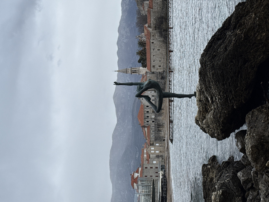 A bronze statue of a ballerina, known as the "Dancer from Budva" or "Ballerina Statue," is centrally positioned on a dark, textured rock formation in the foreground, facing right with her left leg bent and raised behind her, and her left arm extended vertically upwards. The statue stands against the backdrop of the Adriatic Sea, which exhibits choppy, dark grey water under an overcast sky.
In the midground, across the water, lies the Old Town of Budva, Montenegro. The Old Town is characterized by tightly clustered stone buildings with terracotta tiled roofs and ancient defensive walls. A prominent, light-colored bell tower with arched openings, belonging to the Church of St. Ivan, rises above the rooftops on the right side of the Old Town. A sandy beach area is visible along the Old Town's waterfront, with a wooden or metal pier extending into the water. Palm trees are discernible within the Old Town's confines.
Further in the background, a range of dark, rugged mountains stretches horizontally under a uniformly grey, cloudy sky. The overall scene depicts a coastal urban landscape under cool, diffused lighting. No human figures are present apart from the statue.