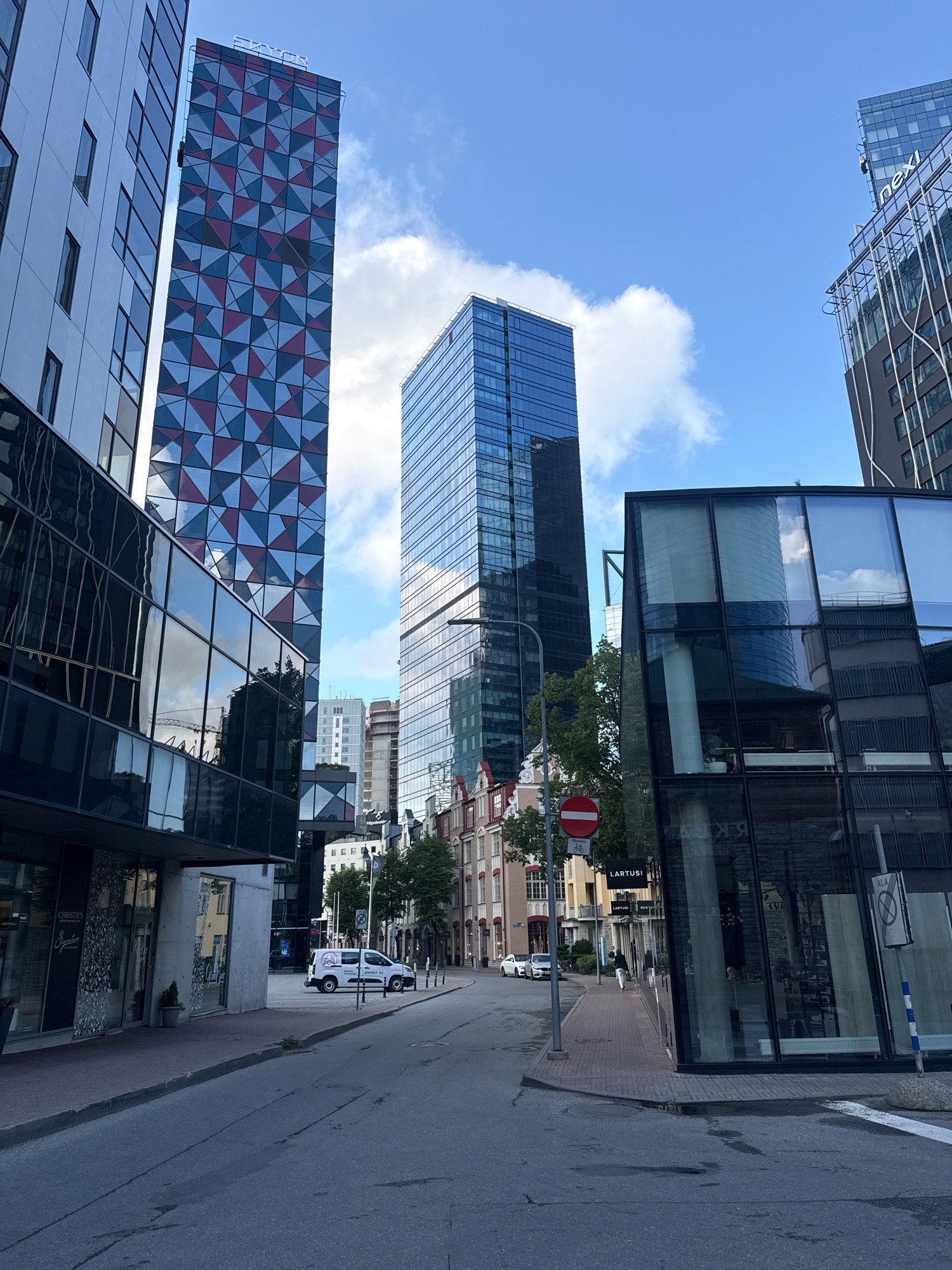 The image captures a street scene in the modern business district of Tallinn, Estonia, specifically the area around Rotermann Quarter and the adjacent newer developments. Tall glass and steel buildings dominate the skyline, showcasing contemporary architecture with reflective surfaces. The tallest building on the left features a distinctive facade with a repeating geometric pattern of red and blue triangles. To its right, a sleek, blue-glass skyscraper rises, reflecting the sky and clouds. In the foreground, a modern building with extensive glass windows is visible, with a street running between it and older, more traditional European-style buildings across the road. A white van with signage is parked on the right side of the street, and a person is walking on the sidewalk. Traffic signs, including a red circular sign with a horizontal bar indicating "No entry" and a sign with a bicycle symbol, are present. Trees and greenery are interspersed between the buildings, softening the urban landscape. The sky is partly cloudy, with a clear blue expanse above. The overall impression is one of a vibrant, evolving city with a blend of historical context and cutting-edge architectural design.