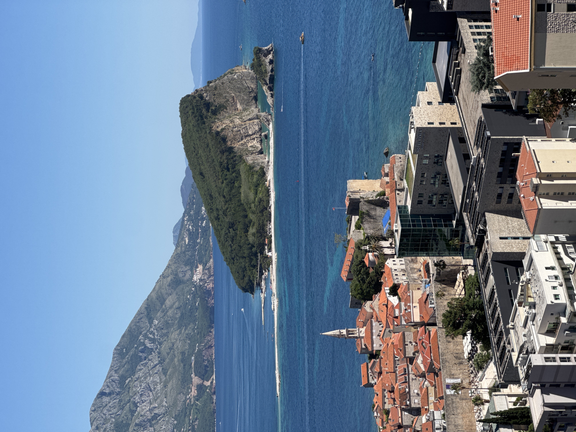 The image captures an elevated view of Budva, Montenegro, focusing on the Adriatic Sea coastline, mountains, and built environment.
The Adriatic Sea occupies a substantial portion of the scene, exhibiting deep blue hues. Several small boats and yachts are scattered across the water. A mountainous landscape rises in the background, its peaks fading into a hazy distance. A smaller island with cliffs and lush green vegetation separates the coastline, and a beach stretches along the shore between the island and the mainland.
The Old Town of Budva is visible on the right side of the image, characterized by closely packed buildings with terracotta rooftops. A church steeple punctuates the skyline. Modern buildings of varying heights and architectural styles rise behind the Old Town. Vegetation is interspersed among the buildings.
No people are discernible due to the distance.