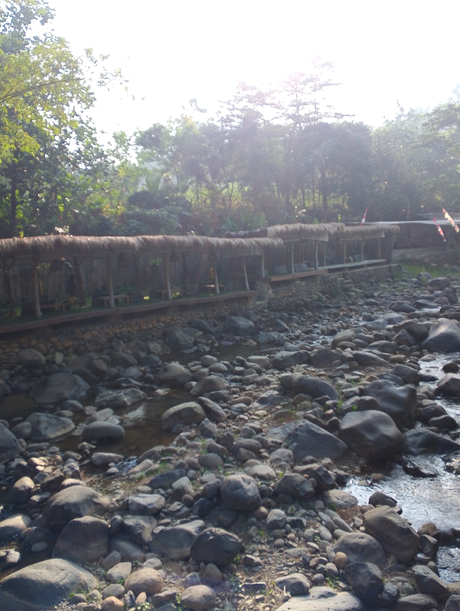 This image depicts a scene in Babakan Madang, Indonesia, featuring a stilted, thatched-roof walkway built over a rocky riverbed. The walkway, supported by wooden posts, appears to house small, open-air seating areas. The riverbed is densely packed with various sizes of grey, smooth stones, with a shallow stream of water flowing between them. Lush green trees and foliage flank the walkway and river. The sun is bright, creating a hazy, overexposed effect in the upper portion of the image and casting dappled light on the scene. Red and white flags are visible on the right side of the walkway, and possibly on the roofline. There are no people visible in the image.