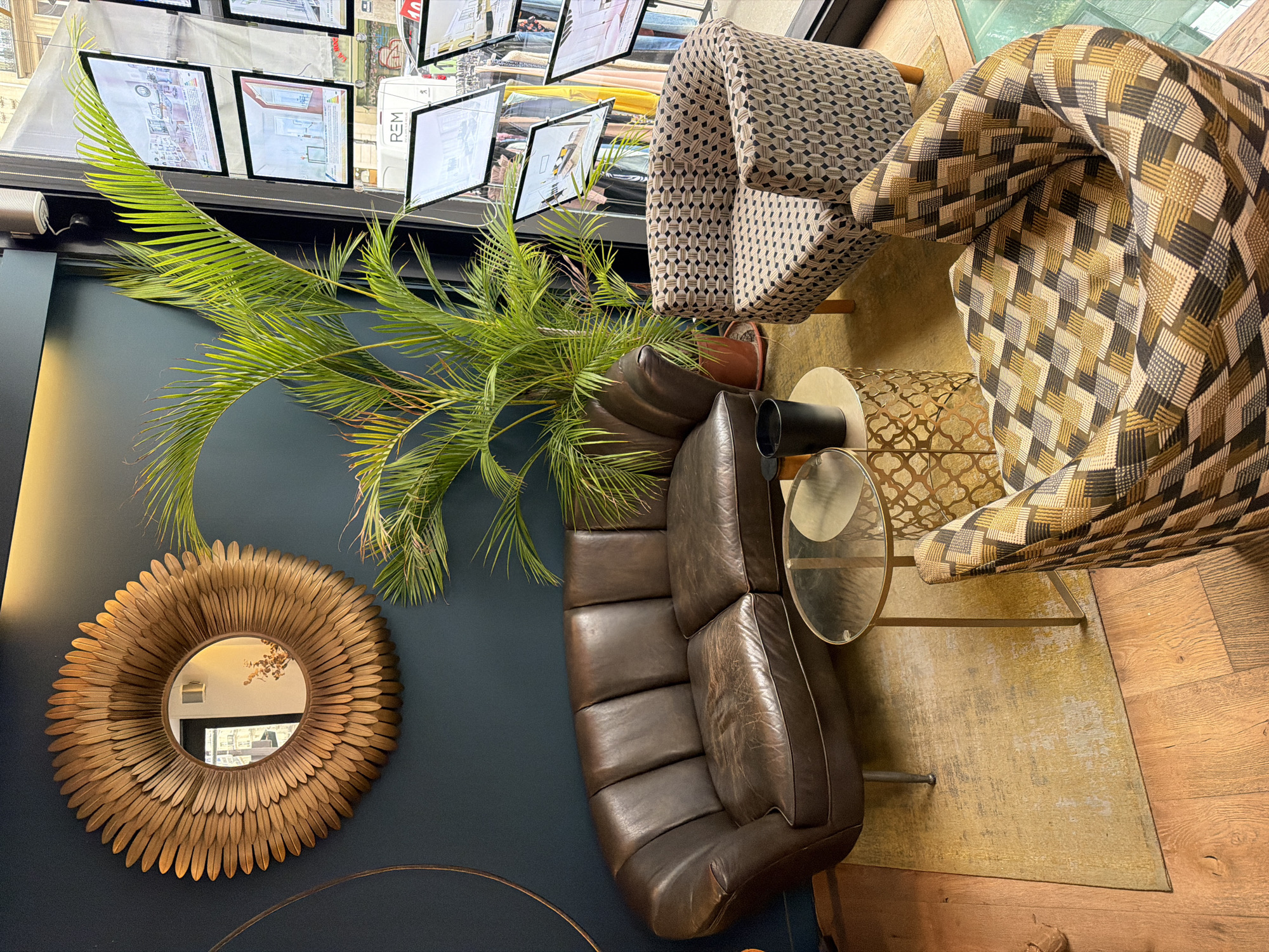 The image is an overhead shot of a room with a contemporary interior design. The scene includes a brown leather sofa, two patterned armchairs, a round coffee table, and a large palm plant. A circular, ornate mirror hangs on a deep blue wall. Illuminated real estate advertisements are visible near the ceiling, suggesting the location is likely a real estate agency. Based on the visible "REM" logo on one of the advertisements, and the overall aesthetic, it is likely this scene is located in Paris, France. The floor is made of wood and covered with a yellow rug.