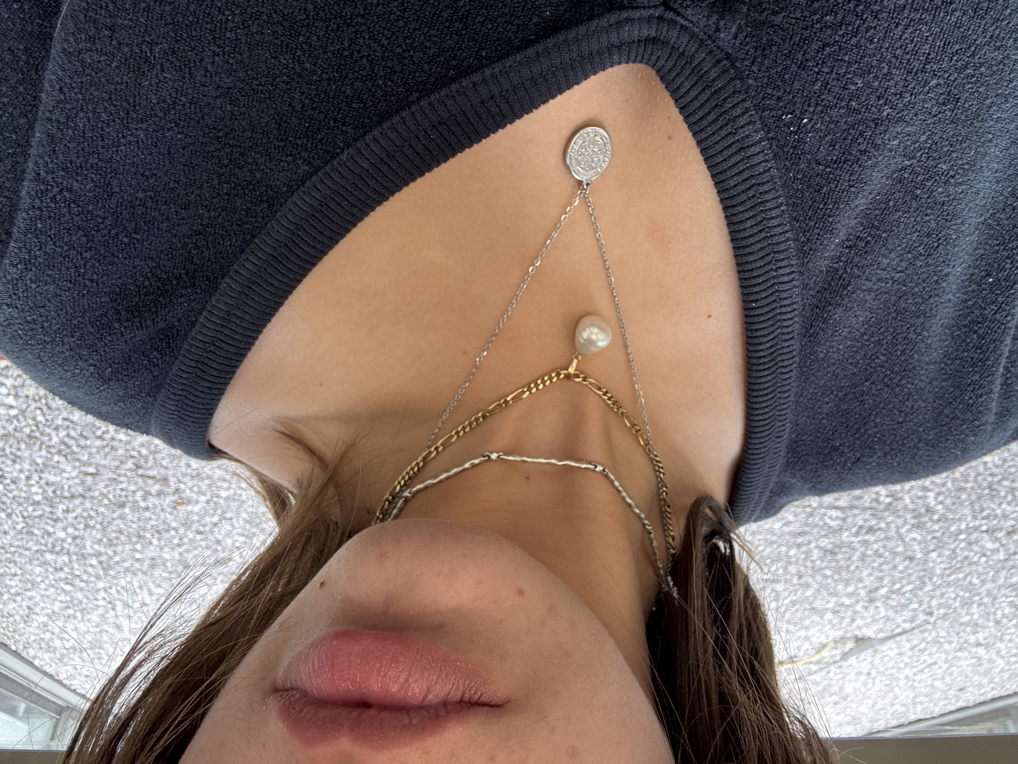 A close-up, inverted perspective captures a person's upper chest, neck, and the lower part of their face. They are wearing a dark-colored, possibly black or navy blue, sweater with a ribbed neckline. Several necklaces adorn their neck, including a multi-layered chain with a decorative coin-like pendant at the top and a pearl-like bead dangling from a middle chain. The person has light skin and pink lips. Brown hair falls across their face and neck. The background, visible above the head, appears to be a textured, light-colored surface or possibly an exterior view through a window. There are no overt indicators in the picture to specifically place it in Huixquilucan de Degollado, Mexico. Therefore, location is unknown.