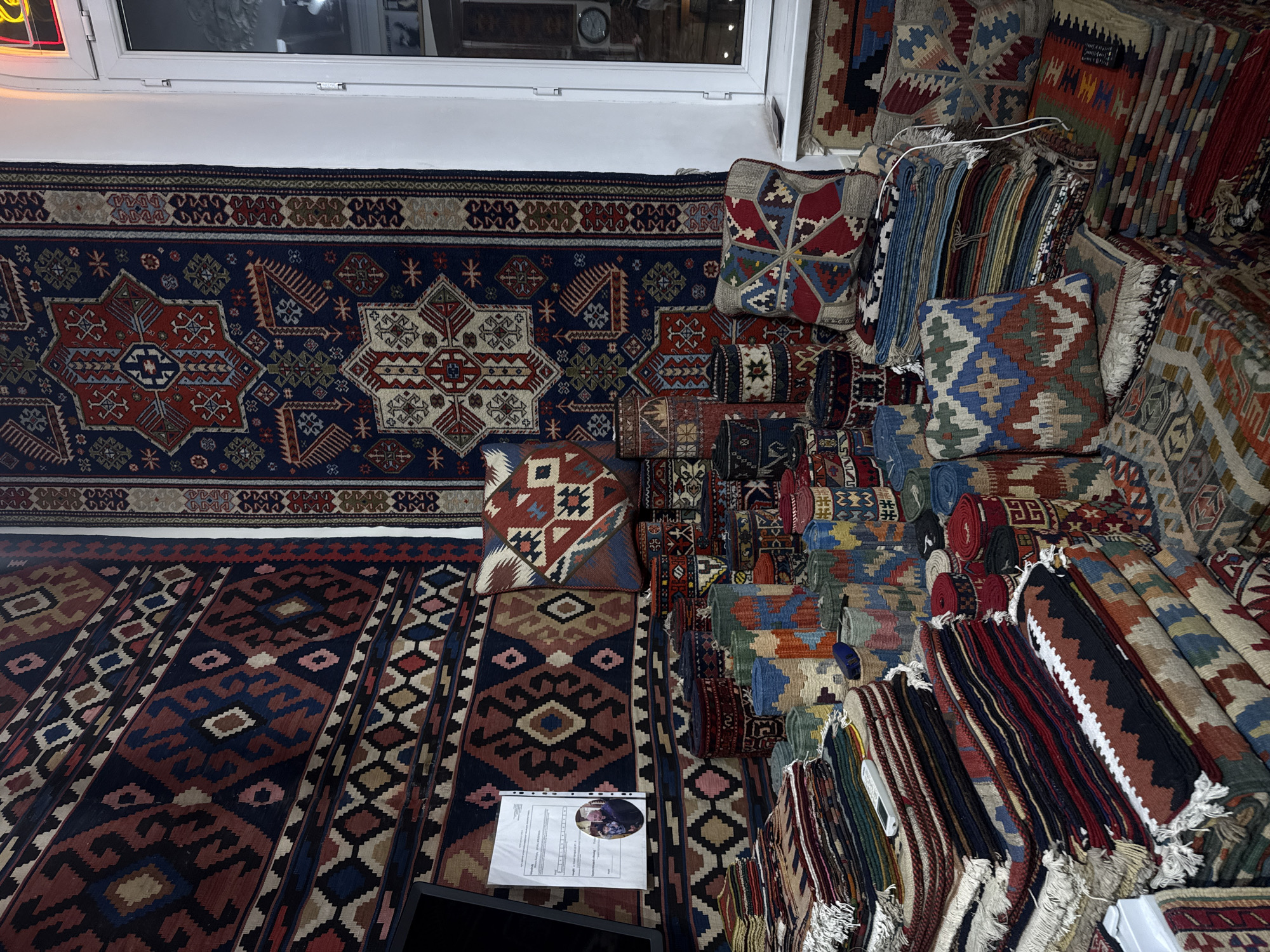 The image depicts an indoor scene in Yerevan, Armenia, featuring a shop displaying various handmade rugs and textiles. The location appears to be a retail space specializing in carpets and related items. The assortment is primarily arranged on the floor and stacked. There is a sign on the rug with an image of a person. The environment suggests a business catering to tourists or residents interested in traditional Armenian crafts.