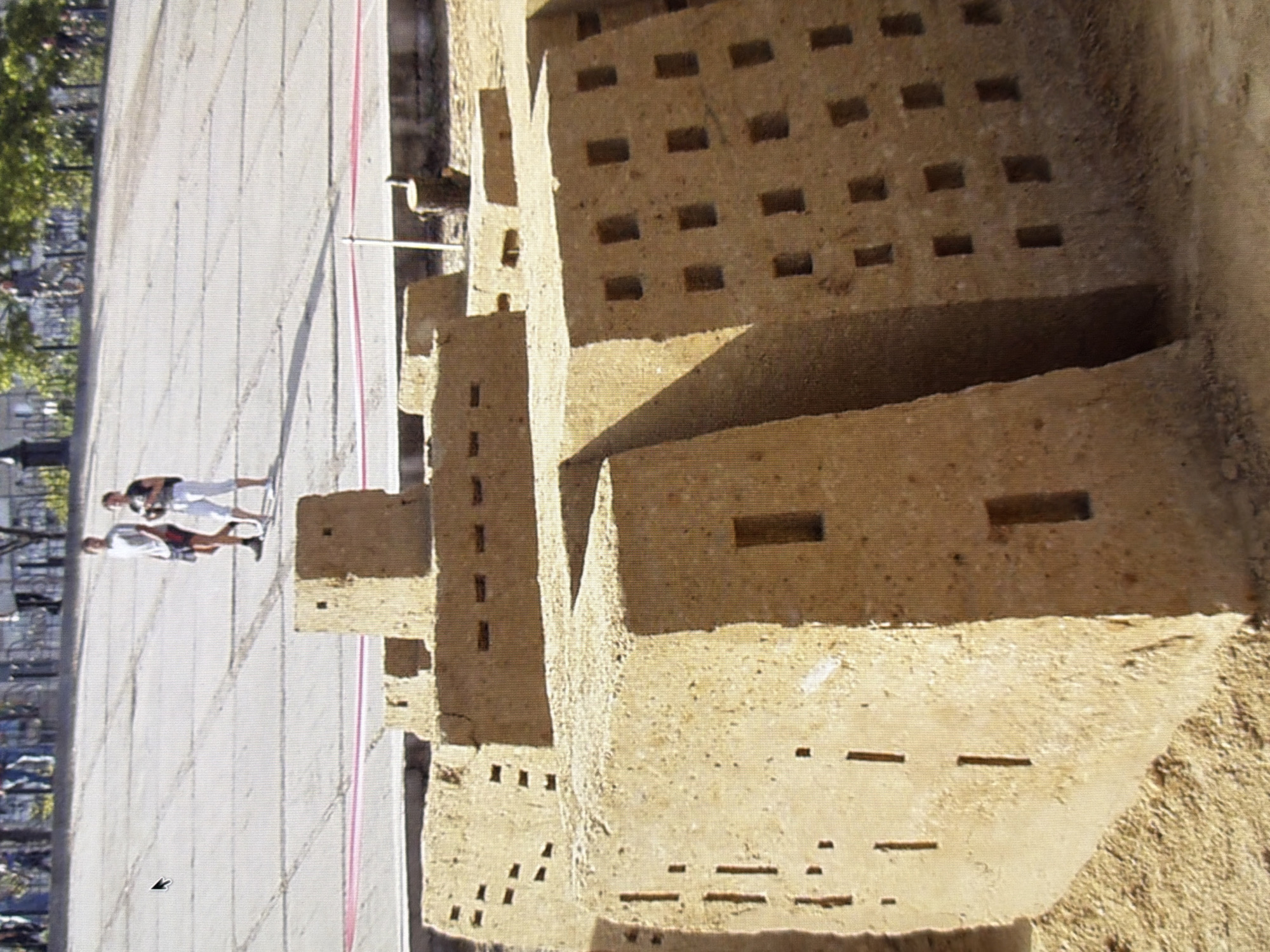 In Kyiv, Ukraine, two individuals walk on a paved surface. In the foreground, a large sand sculpture resembling a fortified structure with numerous small openings is displayed. The sculpture is composed of tan-colored sand and has a textured surface. The environment suggests an outdoor public space with a pathway and some vegetation in the background.