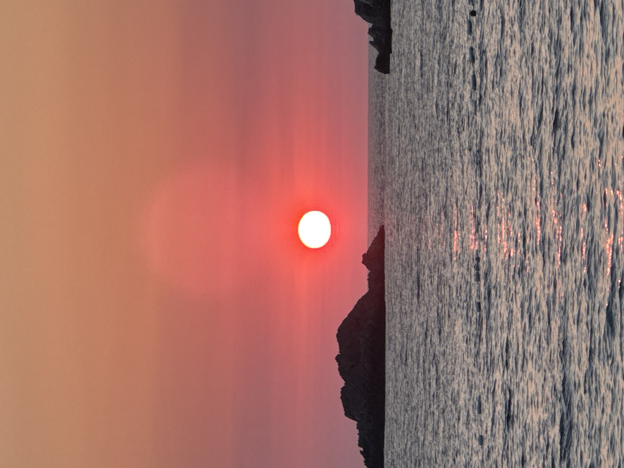 A vertical, eye-level shot captures the serene beauty of a sunset over the Adriatic Sea in Perazića Do, Montenegro. The sky is a gradient of warm oranges and pinks, with the sun, a bright, white orb, positioned slightly right of center, casting a soft glow across the horizon. In the foreground, the calm sea reflects the sky's colors, with subtle ripples disturbing the surface.
Two distinct rocky outcrops emerge from the water. The larger one, a dark, jagged mass, sits to the left of the frame, its silhouette stark against the vibrant sky. A smaller, similarly dark outcrop is visible on the far right, its shape less defined due to the distance. The water itself is a deep blue, with lighter patches where the sunlight hits. A faint line of buoys stretches across the lower right portion of the sea, indicating a contained swimming area. No people or active elements are present, contributing to the tranquil and undisturbed atmosphere of the scene. The overall impression is one of peaceful solitude and natural splendor.