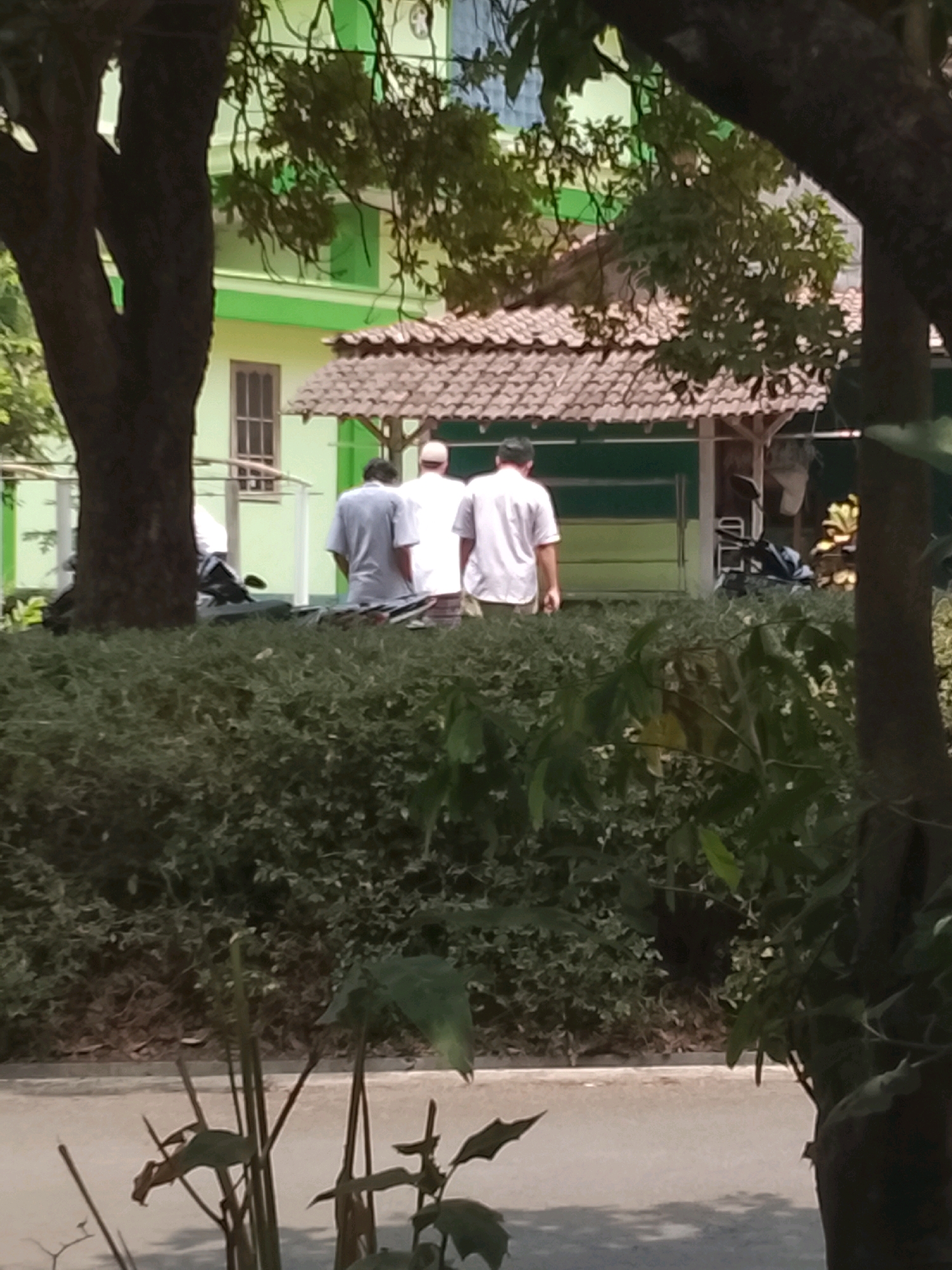 The image depicts a scene at a school or religious institution in Salatiga, Indonesia, characterized by lush greenery and traditional architecture. In the foreground, a dense hedge forms a natural barrier. Behind the hedge, three individuals are visible, facing away from the camera. Two are wearing white shirts and the third is in a grey shirt. They appear to be standing near a building with a tiled roof, which is typical of Indonesian design. The building's exterior is painted a light green, and a window with dark framing is visible. A portion of a tree trunk is on the left side of the image, and another tree trunk is on the right, with foliage partially obscuring the view. A motorcycle is parked near the building. The overall atmosphere suggests a peaceful, outdoor setting, likely during the daytime given the natural light.