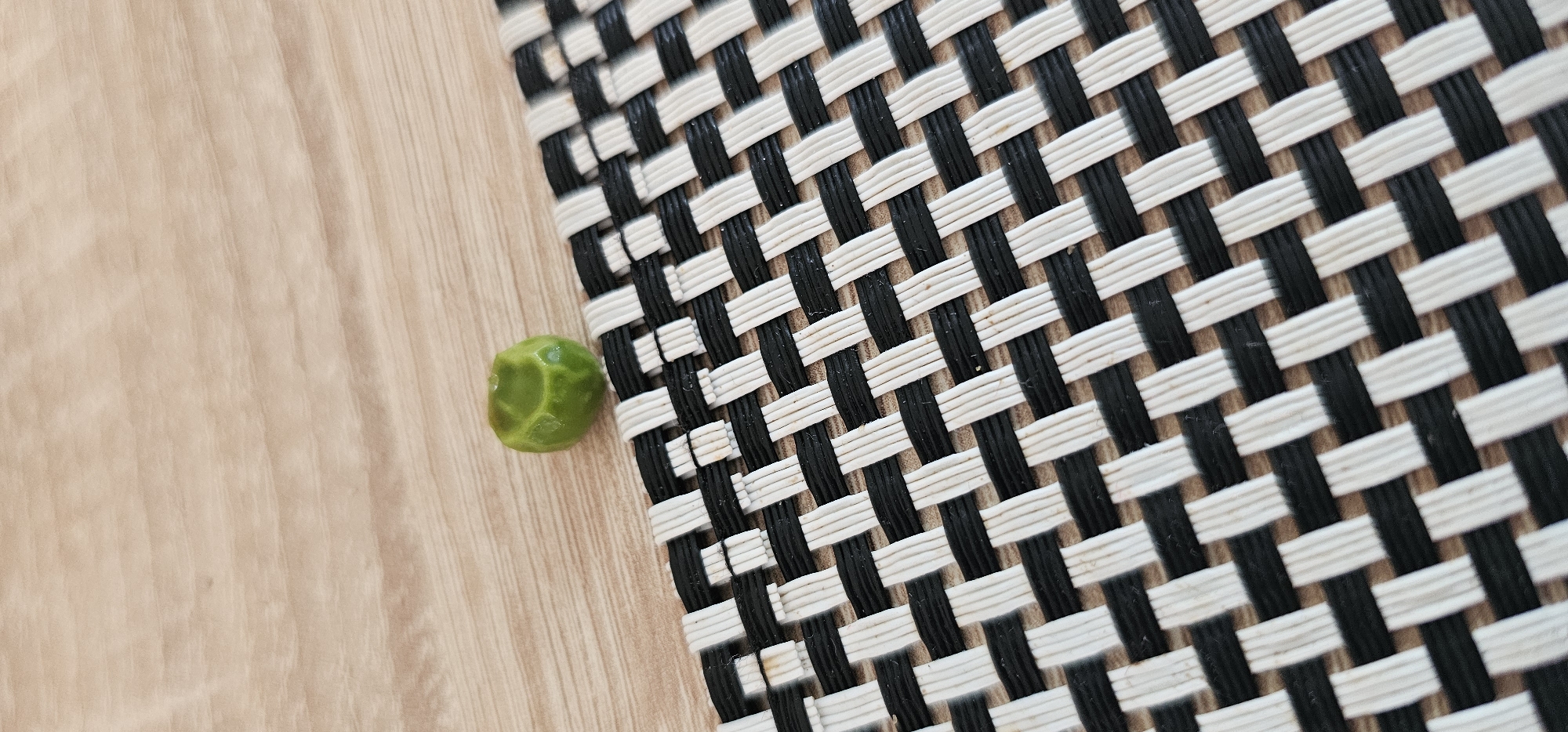 A single green pea sits on a black and white woven placemat. The placemat is positioned on a light brown wooden surface, likely a table. The background is blurred, but the texture of the wood grain is visible. There are no people or discernible actions taking place in the image. The scene is static and focused on the pea and the placemat. The location in the city of Grad Supetar, Croatia, is not discernible from the visual information in the image.