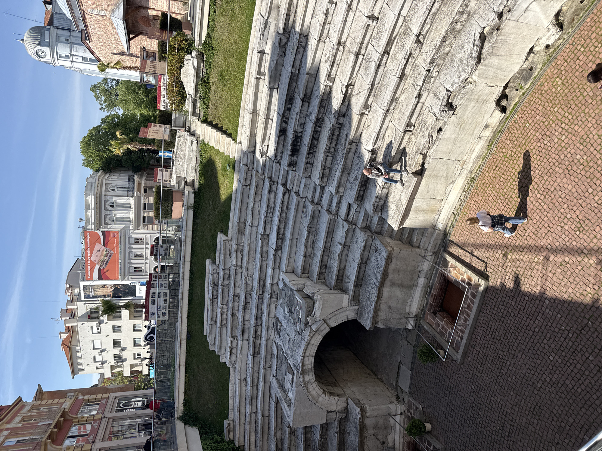 This outdoor image captures the ancient Roman Stadium of Plovdiv, Bulgaria, showcasing its well-preserved stone seating tiers. The stadium occupies the foreground and midground, with its curved stone steps rising towards the left. On these steps, one individual is seated, observing the surroundings. To the right, a paved area of reddish-brown cobblestones stretches out, with another person standing there, casting a long shadow.
Beyond the historical structure, a modern urban landscape unfolds under a clear, bright blue sky. On the far left, an ornate white building featuring a dome-shaped clock tower stands out. Adjacent to it, other buildings of varying architectural styles, from classical to more utilitarian, line the background. Prominently visible are large advertising billboards mounted on these buildings; one displays Bulgarian text, part of which reads "Търсиш обява за ... не губи време..." (Looking for an ad for... don't waste time...) and features the "Samsung" logo. Green trees and foliage, including a palm tree, are scattered among the buildings and alongside the ancient stadium.
The scene is set during daylight hours, indicated by the strong sunlight and distinct shadows cast across the stone steps and pavement, suggesting clear weather. The image captures a peaceful moment at a historical site that is deeply integrated into a contemporary city environment.