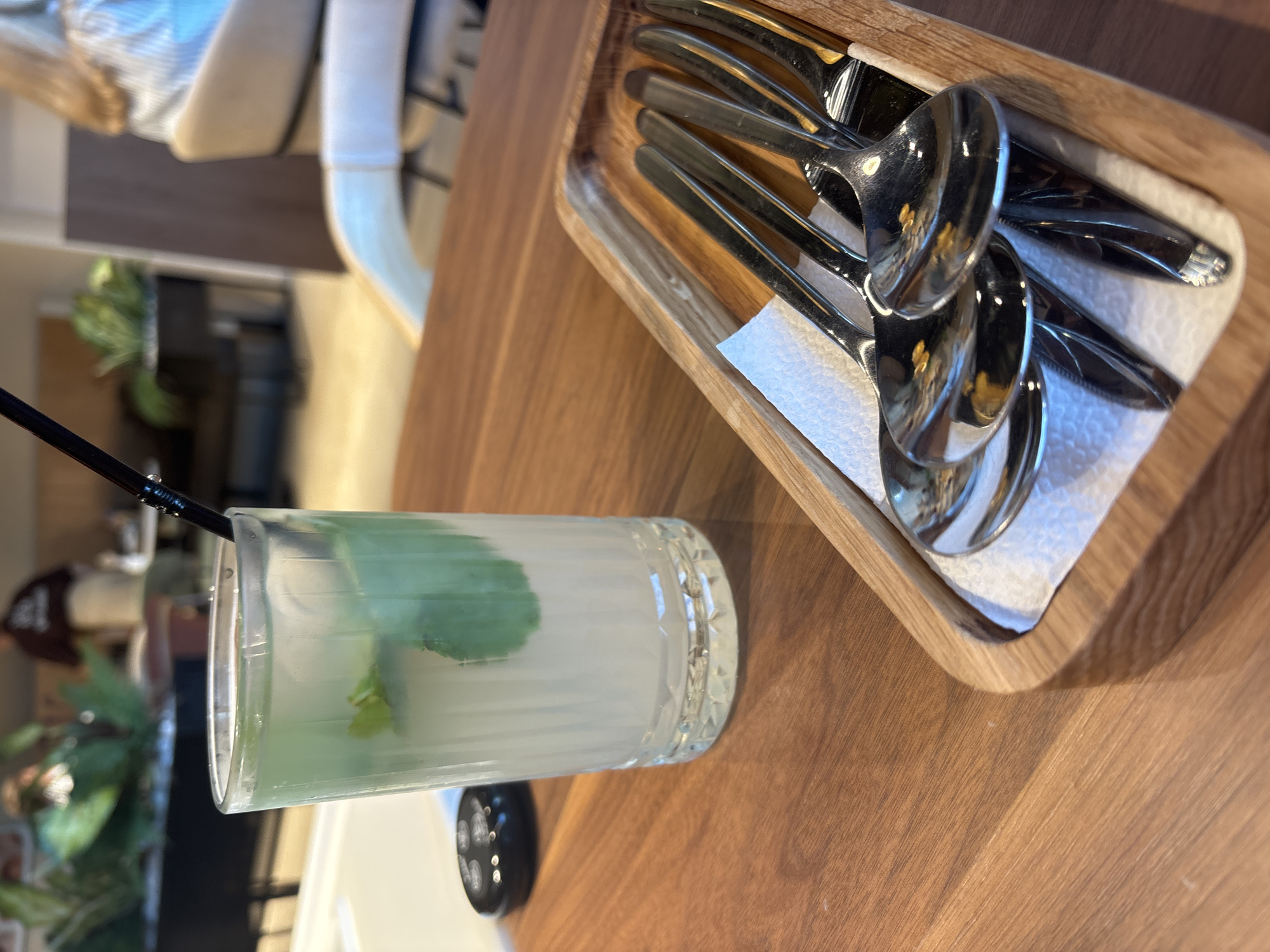 In a bright, indoor setting, likely a cafe in Naberezhnye Chelny, Russia, a clear glass filled with a refreshing beverage and mint leaves sits on a polished wooden table. A black straw protrudes from the drink. To the right, a wooden tray holds several shiny metal spoons and forks, arranged neatly on a white napkin. In the blurred background, diners and indoor plants are visible, hinting at a relaxed ambiance. The scene is captured from a close-up perspective, focusing on the drink and cutlery.