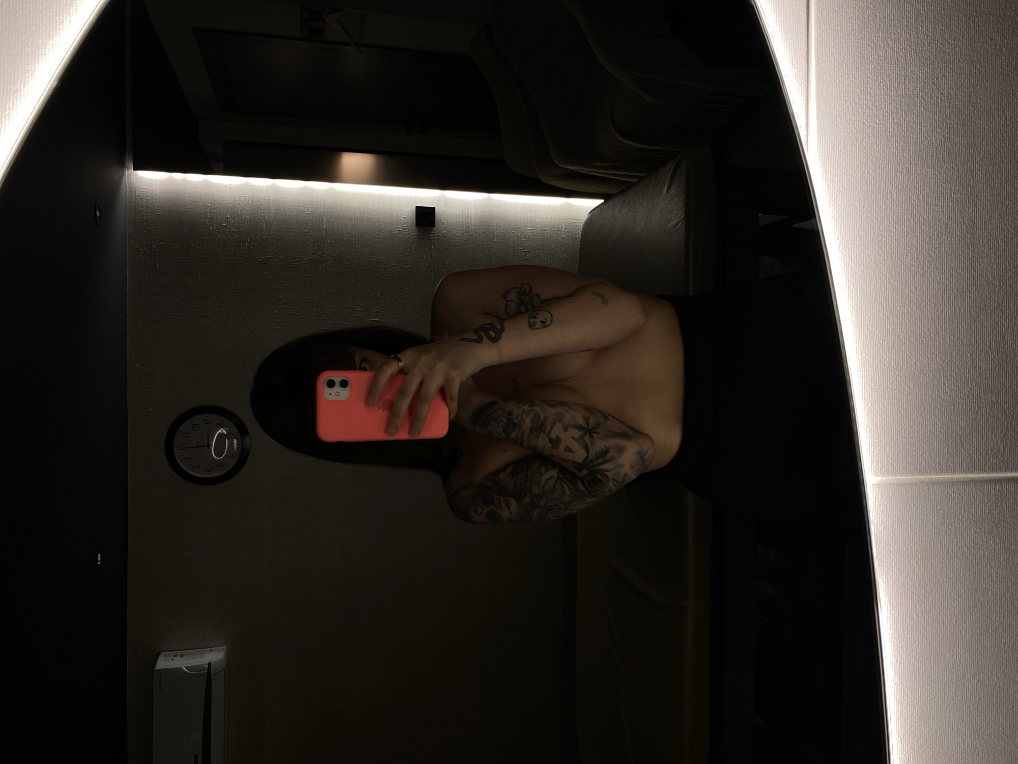 An intimate mirror selfie captures a person in a dimly lit indoor setting, possibly a private room or dressing area. The subject, who appears to be female with long dark hair, is holding a vibrant pink smartphone to take the picture, partially obscuring her face. She is bare-chested or topless, showcasing numerous detailed tattoos covering her arms and shoulders, including a snake, a skull, and a flower, among other intricate designs.
The environment is characterized by dark walls and striking, bright LED strip lighting that delineates architectural features, illuminating the top edge of a dark panel and a textured white wall on the right. In the background of the reflection, glimpses of what appears to be light-colored, padded seating, like a sofa or bench, can be seen. On the dark wall to the left, a circular wall clock with a dark face and white numerals (parts of 10, 11, 12, 1, 2, 3, 4, 5 are visible, with a white circular outline in the center) is mounted. Below the clock, a small, light-colored wall-mounted object is partially visible. The overall mood is dark and modern, with the strong artificial light highlighting textures and creating a dramatic contrast. No specific external cues indicate the provided location context of Votkinsk, Russia.