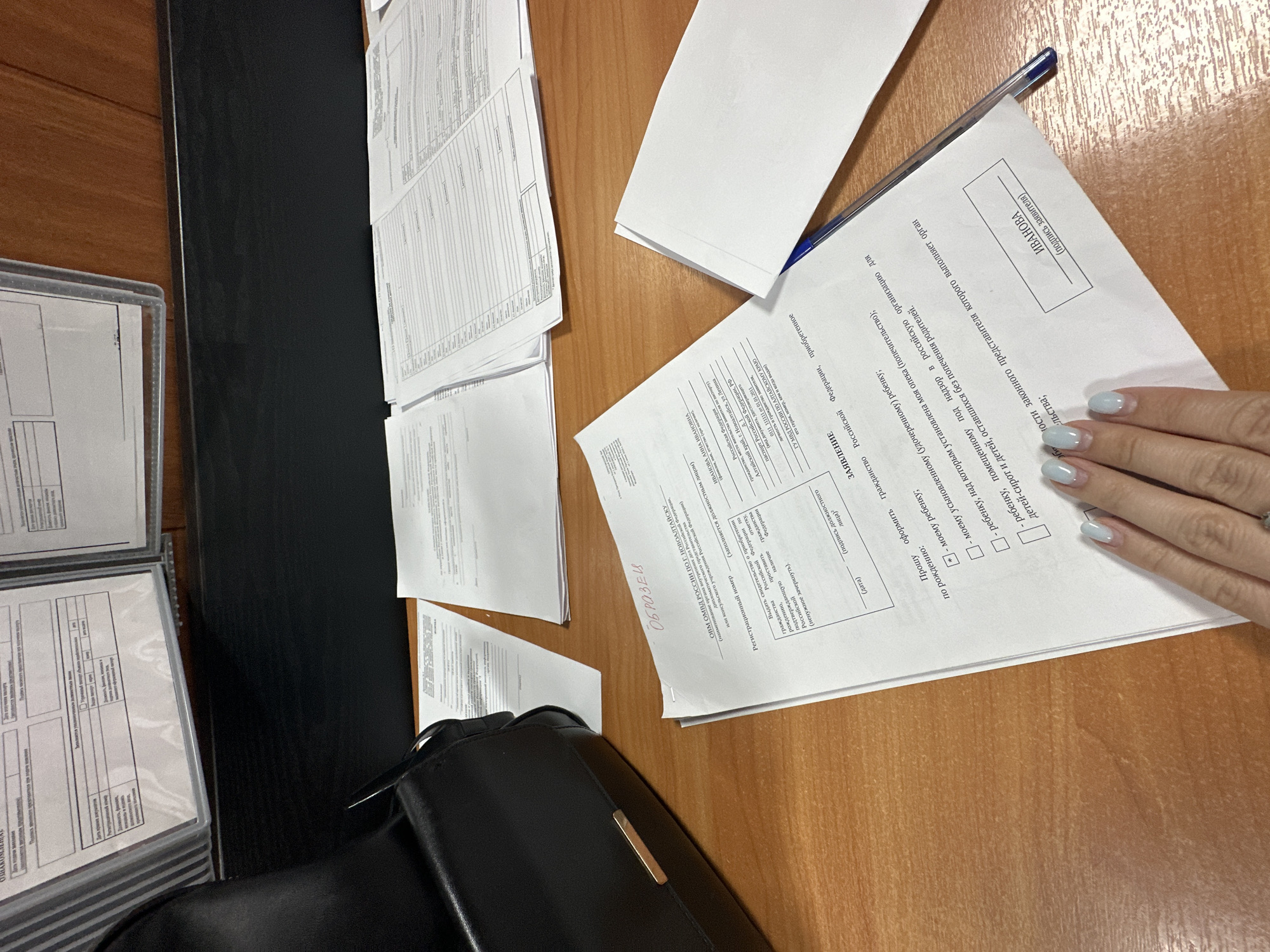 The image displays a close-up, overhead view of a wooden desk surface with several documents and a black handbag. In the foreground, a white document with Russian text is prominently featured, showing a section labeled "ЗАЯВЛЕНИЕ" (Application). Below this, a human hand with light blue nail polish is visible, reaching towards the document. A blue pen rests on the right side of the document.
To the left of the foreground document, a stack of other white documents is visible, some of which are partially obscured. In the upper left corner, a portion of a black handbag with a gold clasp is present. In the background, there are more documents laid out on the desk and what appears to be a wall with framed documents.
The document in the foreground contains handwritten and printed Russian text, including fields for registration number, names, and declarations. Notably, the header of this document indicates "ОВИМ ОМВД РОССИИ ПО Г. НОВОАЛТАЙСКУ" (Migration Department of the Ministry of Internal Affairs of Russia for the city of Novoaltaysk), placing the scene within the city of Novoaltaysk, Russia. Other text on the document includes "ИВАНОВА" (Ivanova), likely a name, and "гражданство Российской Федерации" (citizenship of the Russian Federation). The document also mentions passport details and the possibility of adoption or guardianship of a child.