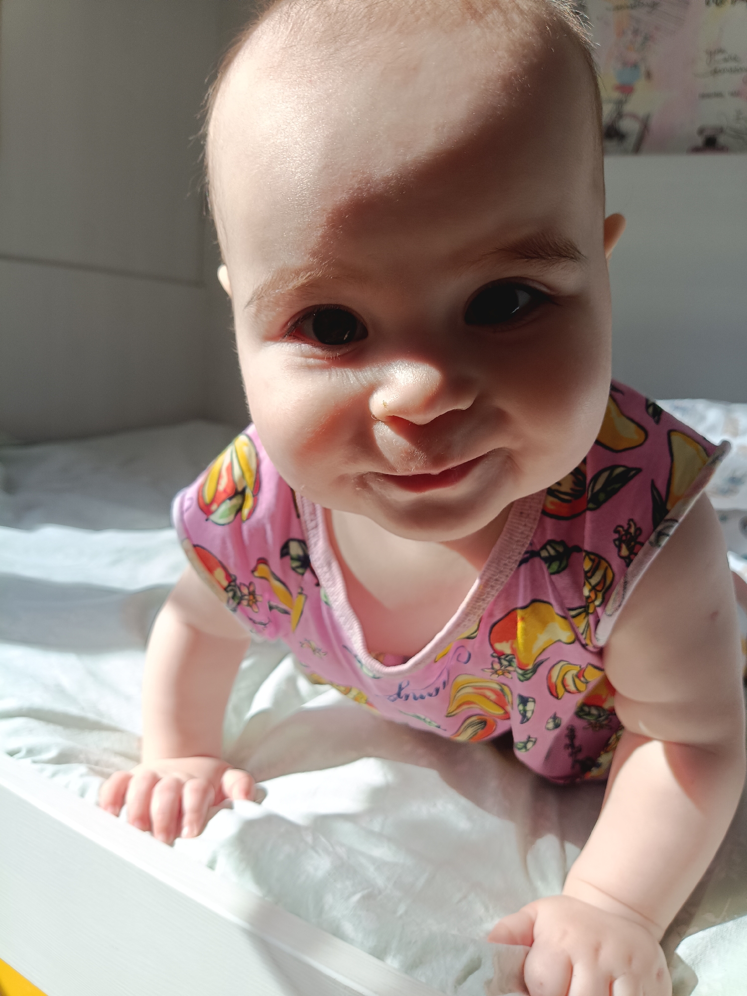 A baby, likely between 6 and 12 months old, is positioned on their hands and knees on a white, possibly cotton, bed sheet. The baby wears a pink sleeveless shirt with fruit-patterned illustrations in shades of yellow, red, and green. The baby's skin is fair, and they have dark eyes. A portion of a white headboard or crib frame is visible on the lower left. Bright, direct sunlight illuminates the left side of the baby's face and the adjacent area of the bed. The background suggests an interior room, and there's a hint of what might be artwork or papers on a wall behind the baby's head. There's no indication of Noviy, Russia.