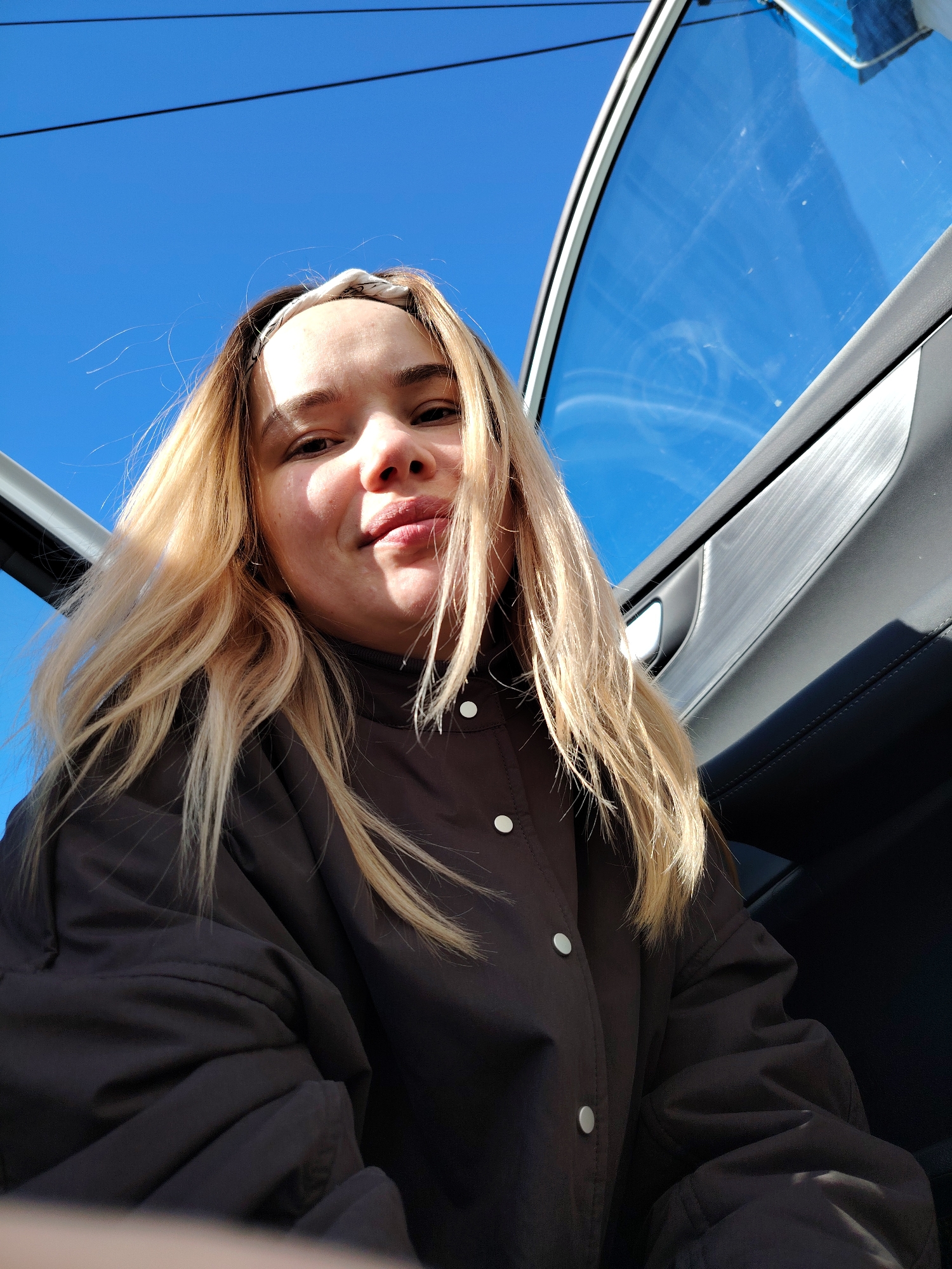 A low-angle close-up shot of a fair-skinned woman with blonde hair wearing a brown jacket with white buttons. She is inside a car with the door open, showing a bright blue sky with a few wispy clouds visible through the open window. The car's interior is black, and the door frame is silver. The location is likely Chaikovsky, Russia.