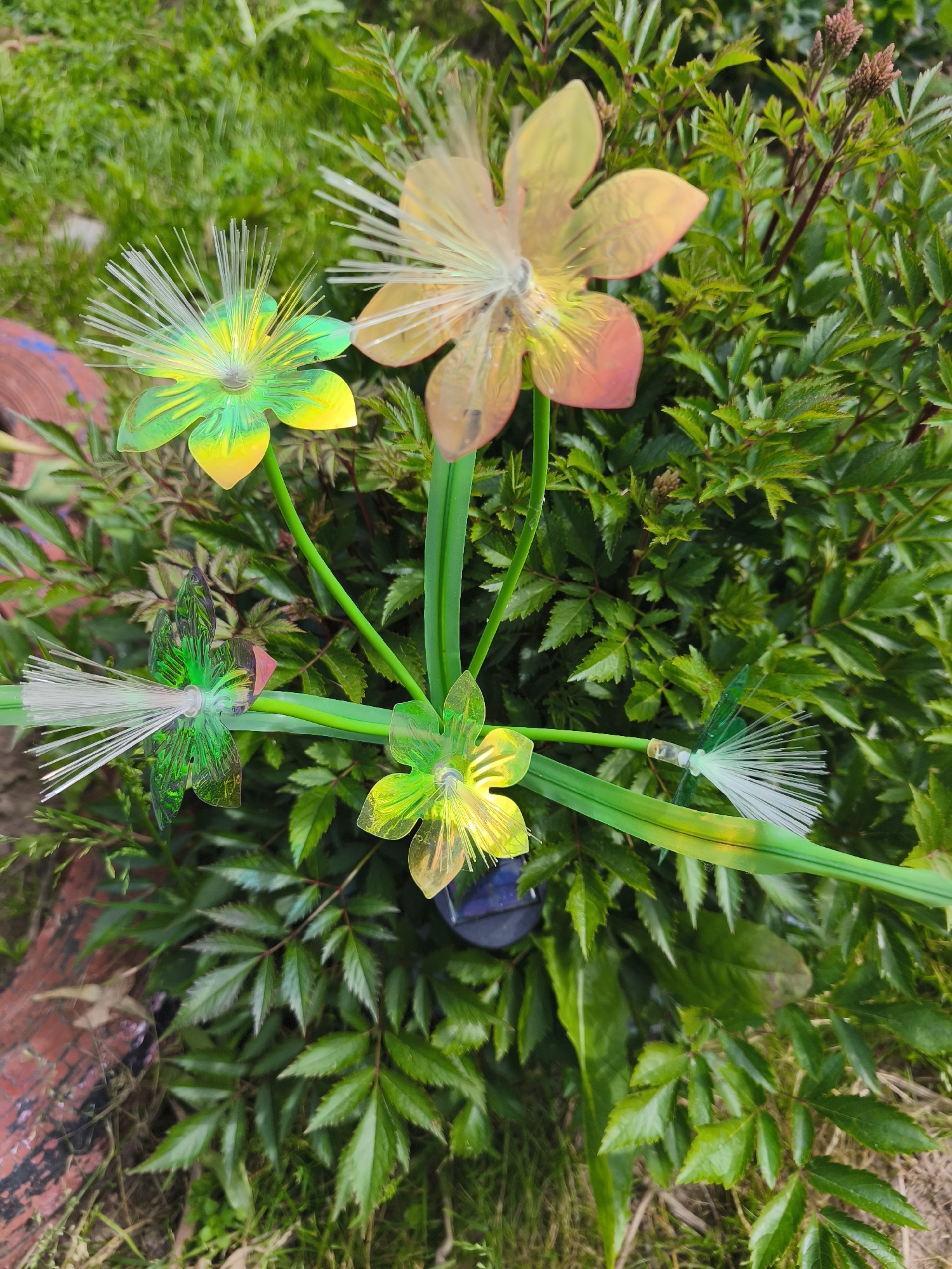 This image depicts a close-up, overhead view of decorative garden ornaments resembling flowers and dragonflies amidst lush green foliage. The ornaments are made of translucent plastic and metal, with bright green and yellow hues, and some feature white, string-like stamens or dragonfly tails. One larger, translucent orange and yellow flower ornament is partially visible at the top right. A dark blue solar panel is nestled amongst the leaves. The background consists of dense, green leafy plants, with hints of dried leaves and what appears to be a red-brown object, possibly a brick or painted stone, on the left. The setting is outdoors, in a garden environment. The location is identified as Puchezh, Russia.