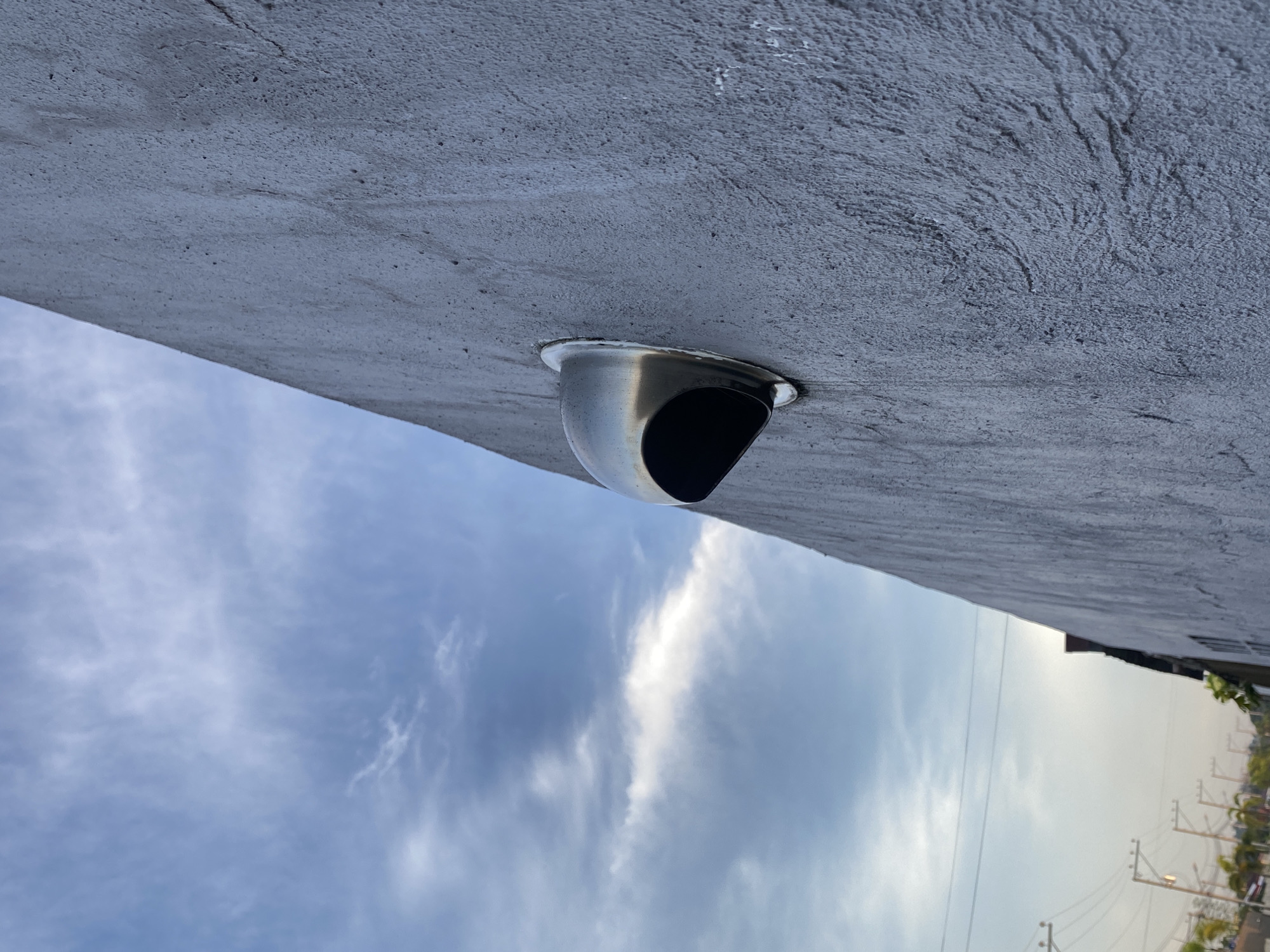 A low-angle, eye-level shot shows a curved, stainless steel vent protruding from a gray concrete surface, possibly a building's overhang or soffit. The vent has a rounded shape with a dark, angled opening. Behind the concrete and vent is a view of the sky, displaying a mix of light blue sky and scattered white clouds. In the distance, some trees, buildings, and utility poles are visible. This image was taken in Davao City, Philippines.
