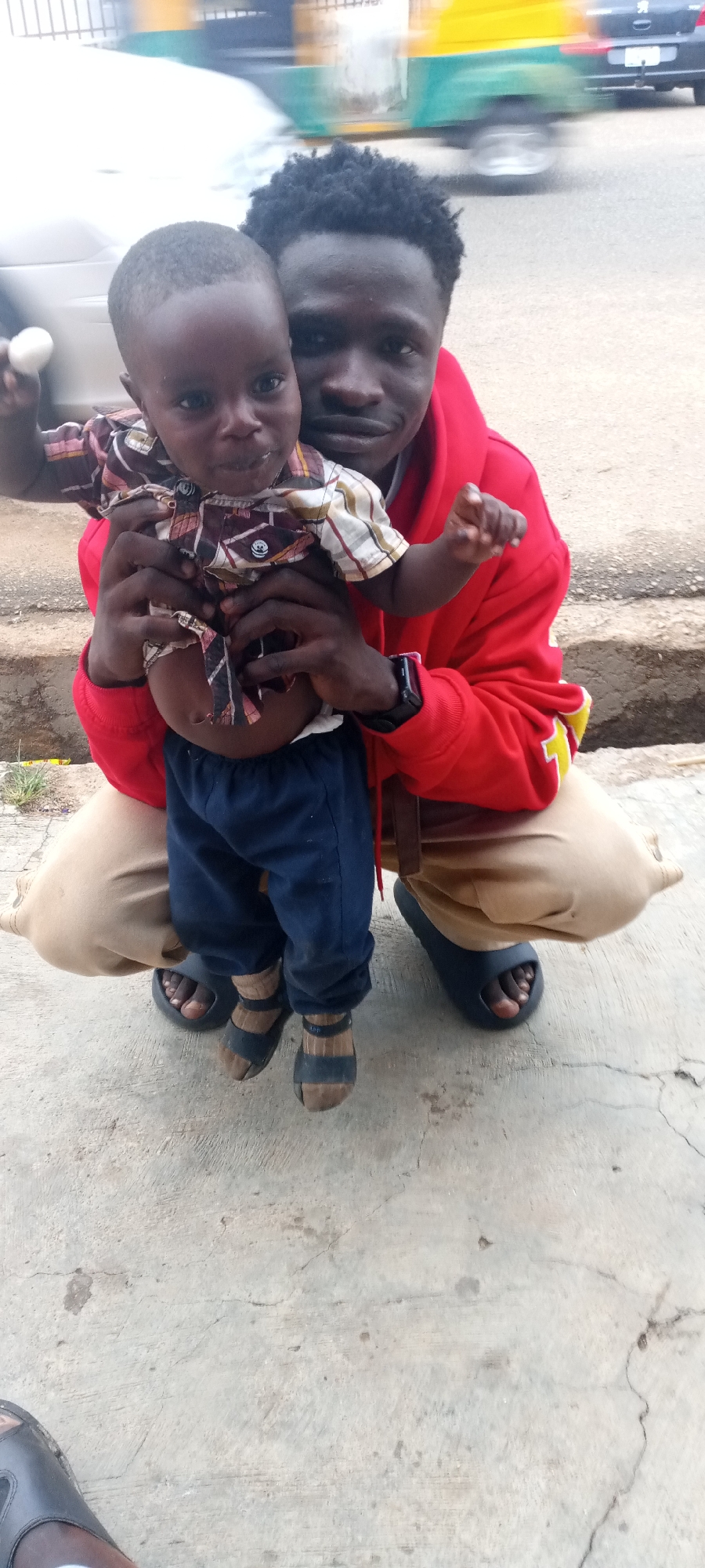 In Jos, Nigeria, an African man in a red hoodie and beige pants is squatting on a concrete surface while holding a young child. The child wears a plaid shirt and blue pants, with socks and sandals. Both are looking at the camera. A street with moving vehicles is visible in the background. The setting appears to be outdoors, likely a roadside or walkway.