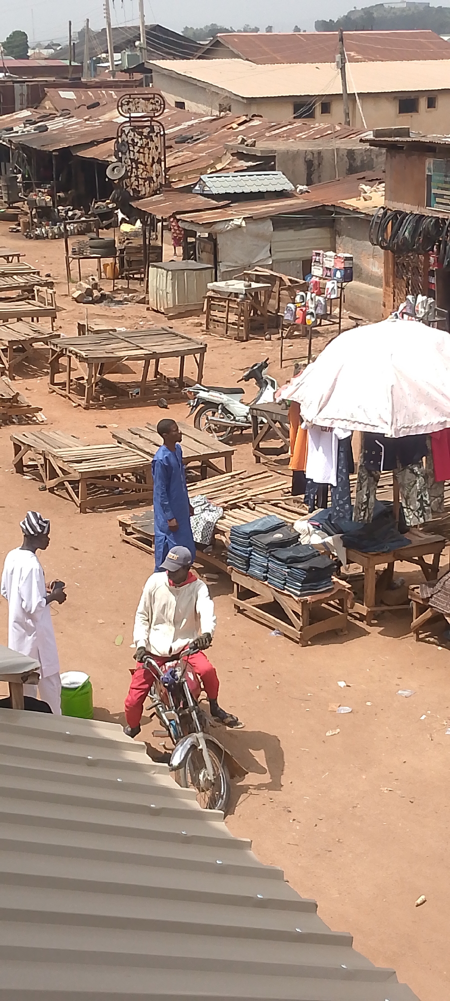 The image depicts an outdoor marketplace in Bukuru, Nigeria. The scene is characterized by a dusty, reddish-brown ground and a variety of stalls constructed from wood and corrugated metal. Many stalls appear empty or are being prepared for the day's business. There are goods such as clothes, tires, and household items for sale. Three men are in the foreground. One man wears a white outfit and a striped hat and holds a dark object in his hand. Another man in a blue robe stands nearby. A third man in a white sweatshirt and red pants sits on a motorcycle. The architecture consists of simple, makeshift structures. Buildings are visible in the background.
