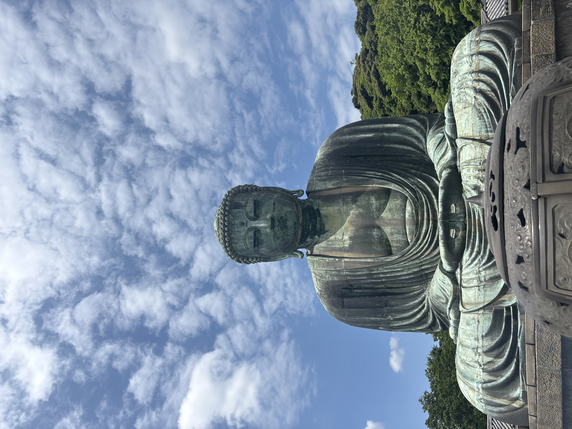 The image shows the Great Buddha of Kamakura (Kotoku-in), a monumental outdoor bronze statue of the Buddha located in the city of Kamakura, Japan. The statue is depicted from a low angle, facing the viewer. The Buddha is seated in a meditative pose, with its hands resting in its lap. The statue is green with age, showing patina. Behind the Buddha, a lush green hillside with trees is visible, and above that, a blue sky with scattered white clouds. In the foreground, part of a large bronze incense burner is visible. The overall atmosphere is serene and majestic, highlighting the grandeur of the statue against the natural backdrop.