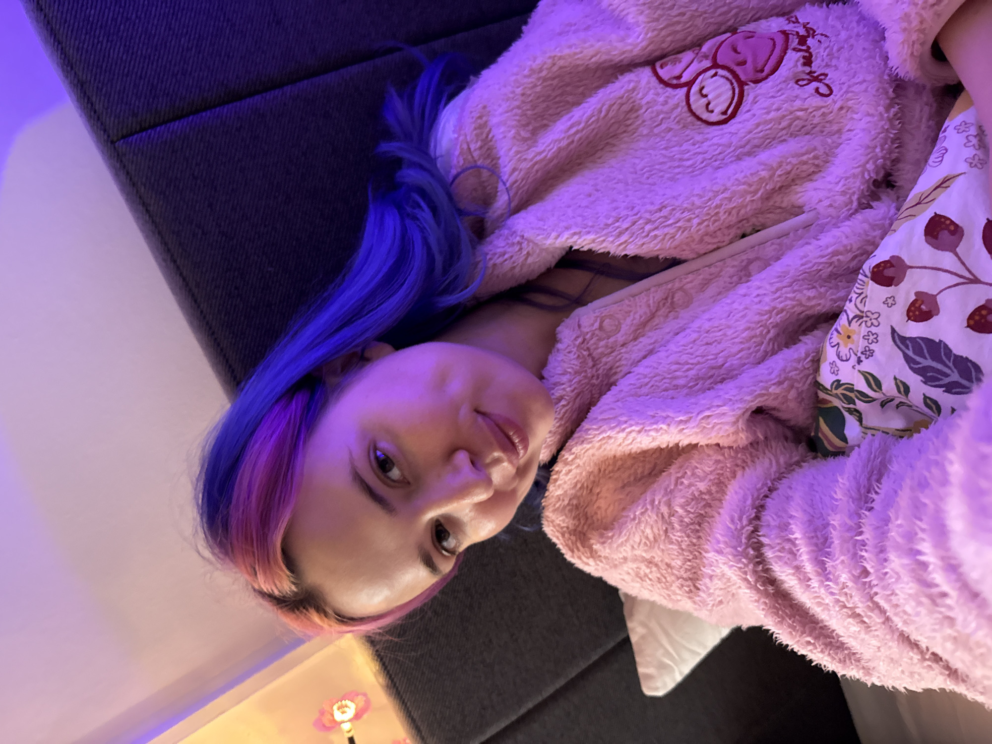 A young woman is captured in a portrait-oriented photograph, lying down indoors, likely in a bedroom. Her head rests on a dark grey, fabric-covered surface, possibly a headboard or bed frame. She has strikingly vibrant hair, dyed in shades of electric blue and pink/purple, which cascades around her head. The woman is wearing a plush, pink, fuzzy robe or blanket, detailed with an embroidered design on the left chest that features two cartoon-like characters. A section of a white sheet or pillowcase with a pattern of red berries and green leaves is visible beside her on the right.
The woman gazes directly into the camera with a calm, slightly smiling expression. The scene is illuminated by soft, predominantly purple ambient lighting, which casts a distinctive glow over the entire image. In the lower-left corner, a small, decorative lamp shaped like a flower emits a warmer, soft orange light, providing a contrast to the cool purple hues. The overall ambiance suggests a relaxed evening or nighttime setting. Although no specific location cues are visible within the image itself, this scene is set in the City of Zagreb, Croatia.