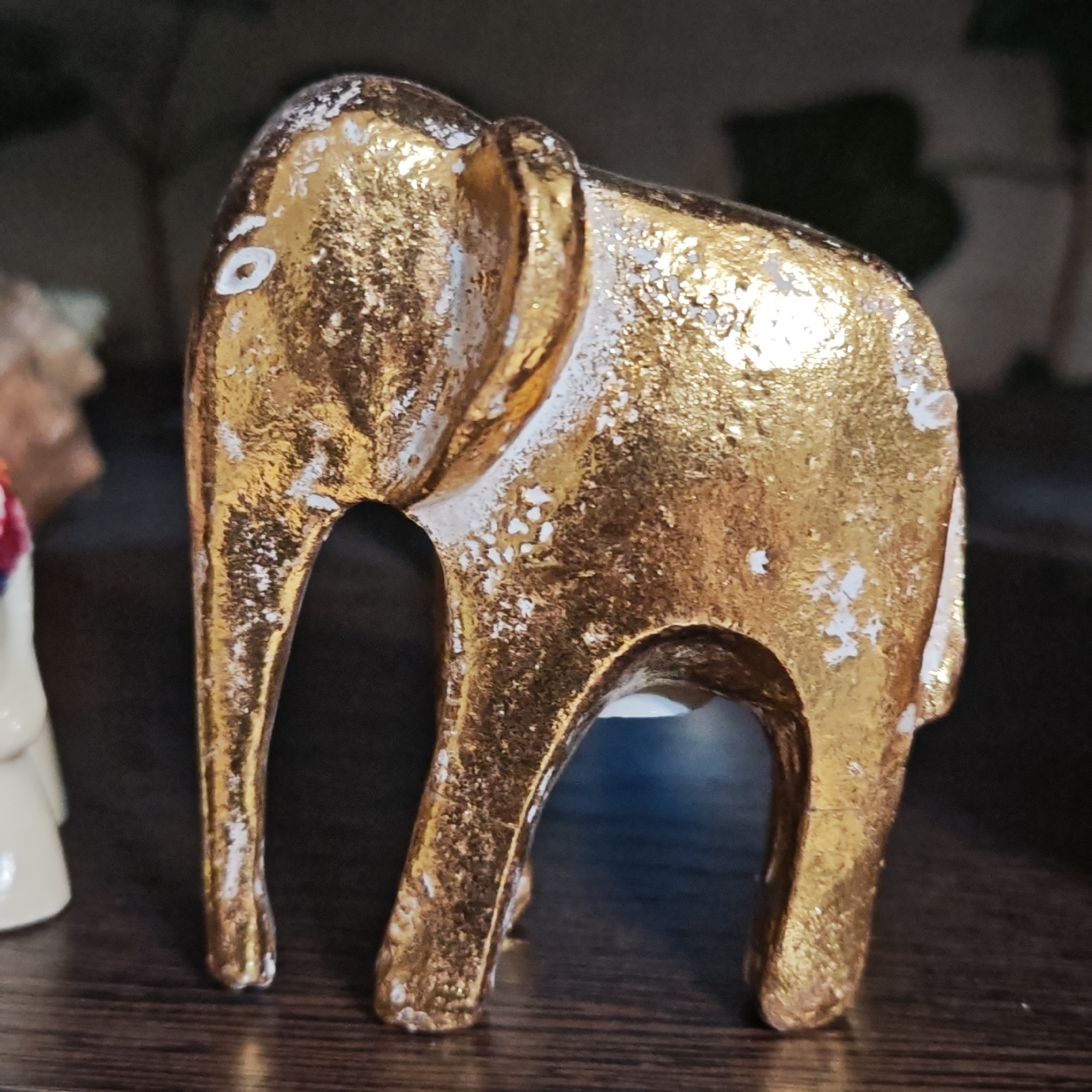 The image captures a detailed shot of a decorative elephant figurine, exhibiting a rich, distressed gold finish with patches of white showing through, suggesting age or an artistic antiqued effect. The small statue depicts a stylized elephant standing firmly on all four legs, with its trunk gently raised. A distinct white circle on its head may represent an eye.
The figurine rests on a dark, horizontally-grained surface, likely wood, enhancing its metallic sheen. The background is intentionally blurred and dark, providing minimal distraction and focusing attention on the elephant. Hints of other objects, including a light-colored item to the left, suggest it is part of an indoor display or collection on a shelf or table. The lighting emphasizes the texture and golden color of the elephant, creating a sense of depth and artistry. No specific activity or event is depicted, as it is a still life. No text is visible in the frame. While provided with the location context of Novo-Nikol'skoe, Russia, there are no discernible visual cues within the image itself to confirm this geographical location.