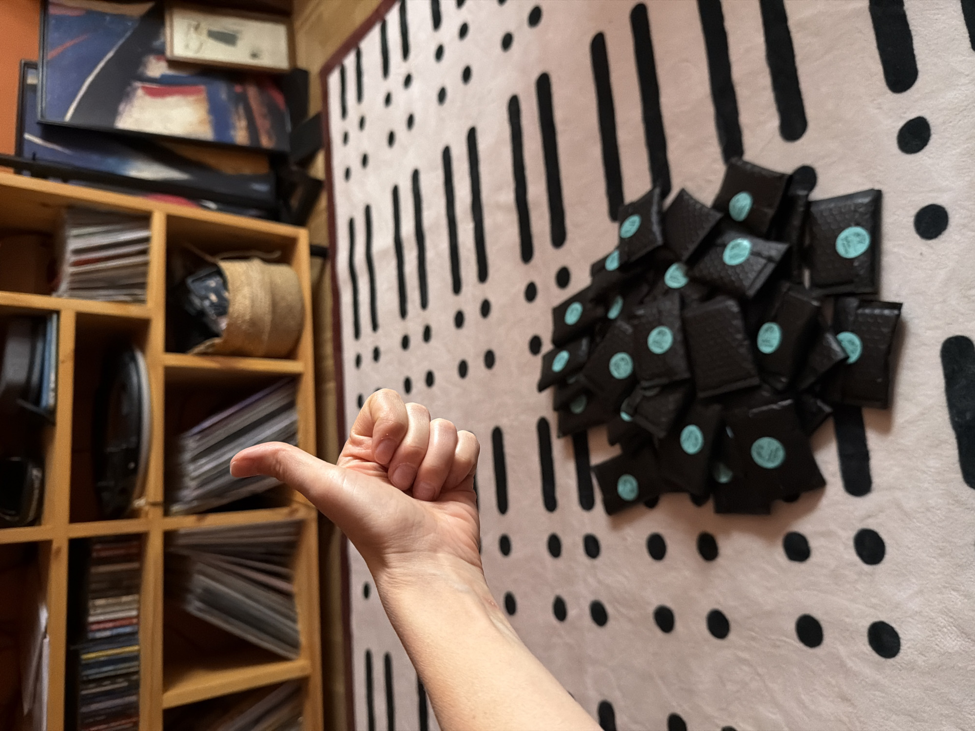 The image captures an indoor scene featuring a hand making a thumbs-up gesture in the foreground. The hand, with light skin tone, extends from the lower left, its thumb pointing upwards.
To the right, a large pile of small, black, rectangular packages or pouches is stacked on a light-colored surface. Each package features a circular, light blue sticker, many of which appear to have a stylized "SM" logo or similar initials. The surface beneath these packages is a textile, possibly a rug or blanket, patterned with numerous black exclamation marks and dots against a light background.
To the left and in the background, a wooden shelving unit is visible, filled with various items. Several shelves hold stacks of CDs or DVDs. One compartment contains a woven, light-brown basket. Other shelves hold darker, less discernible objects. Above the shelving unit, several framed artworks or pictures are stacked. One prominent piece is an abstract painting with bold blue, red, and black strokes. A smaller, lighter-colored framed object rests on top of this stack.
The overall scene suggests a personal or work space, possibly where items are prepared for sale or distribution, indicated by the uniform packaging and stickers. The thumbs-up gesture conveys a positive sentiment, such as approval or satisfaction. The lighting appears to be indoor artificial light. No explicit text other than the potential logo on the stickers is clearly visible.