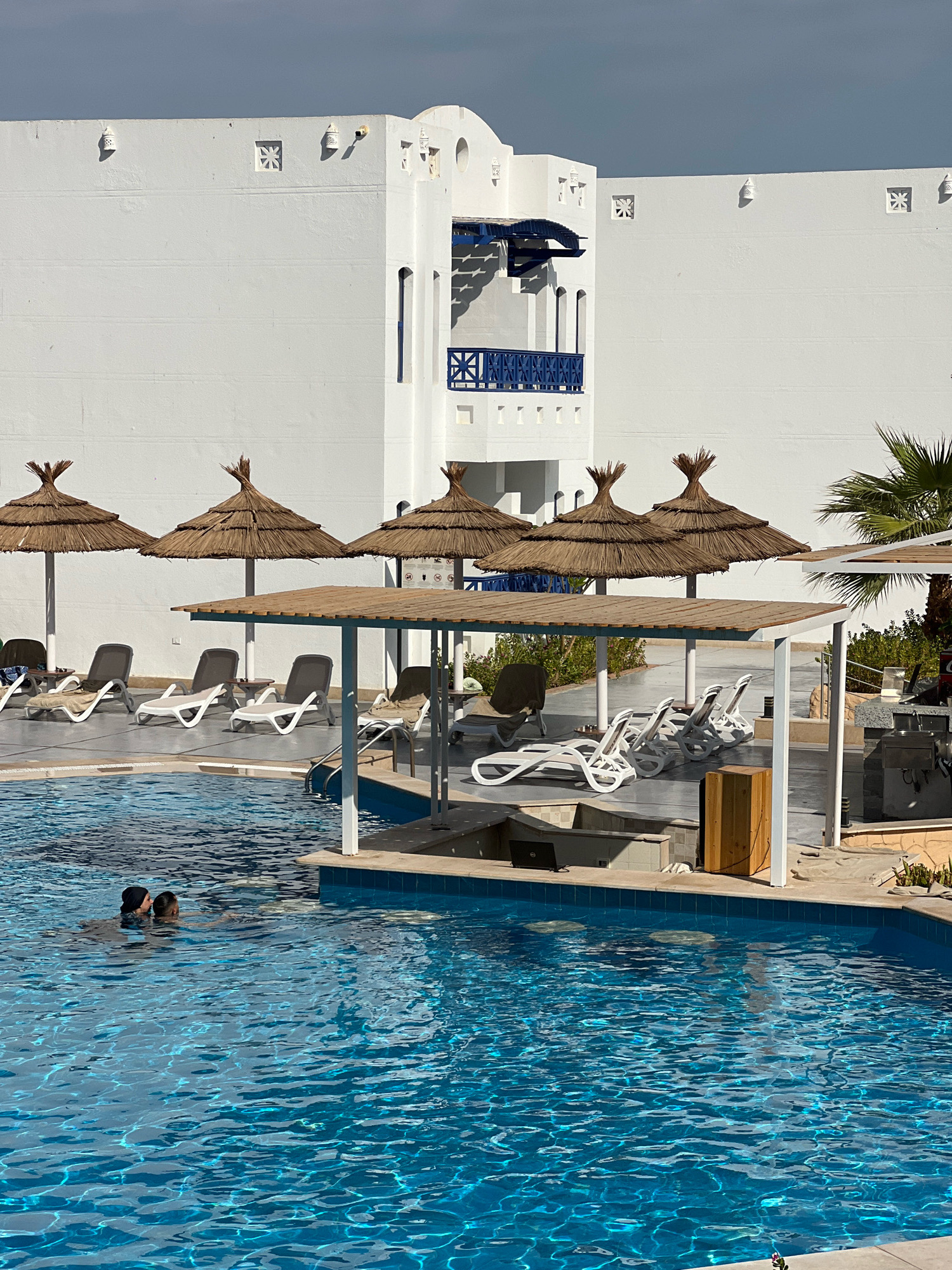 The image depicts a resort swimming pool area in Sharm El Sheikh, Egypt. The pool is a vibrant blue, with two individuals partially submerged in the water. Surrounding the pool are several thatched-roof umbrellas providing shade over rows of lounge chairs. A white building with distinctive blue accents and architectural details, typical of resorts in the region, stands prominently in the background. The scene suggests a sunny day at a leisure facility.