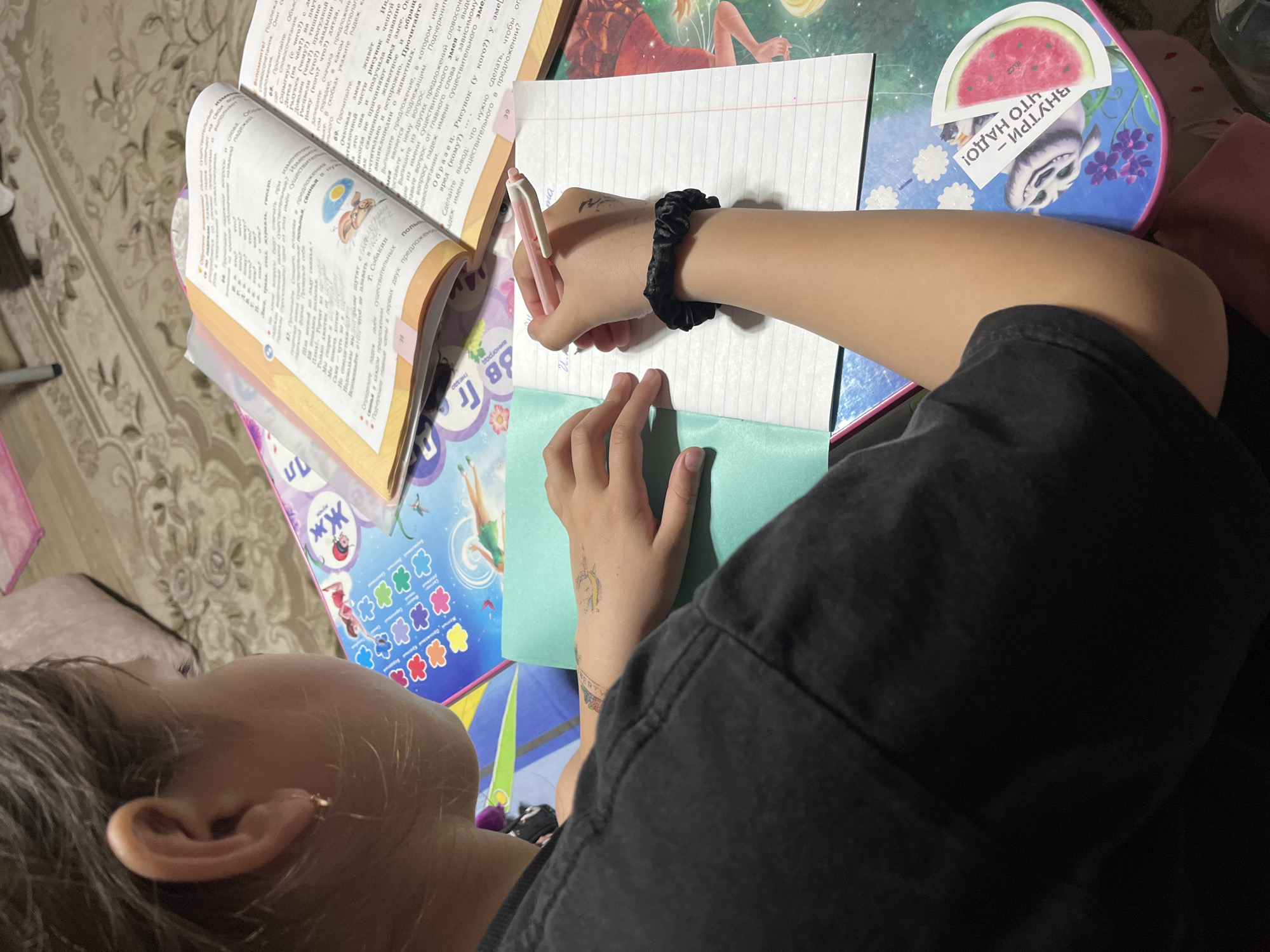 A child is sitting at a table, writing in a notebook. The table is covered with a children's tablecloth and there are various items on it, including an open book with Russian text, a notebook with lined paper, a pink pen, a turquoise envelope, and a small box with a cartoon image. The child is wearing a black shirt and a black scrunchie on their wrist. The child has an earring on their left ear and a tattoo on their left wrist. The photo may have been taken in Novopokrovskaya, Russia, as the book appears to be written in Russian.