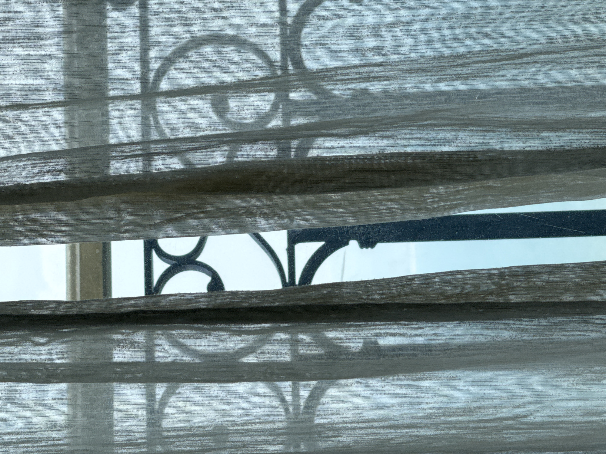 The image presents a view through window blinds, partially obscuring a black metal exterior railing with curved and linear elements. Behind the railing and visible through the blinds and railing's openings is a blurred view of what appears to be a bright sky and possibly buildings. The blinds are horizontal, layered, and textured with a greyish-brown color. There is no evidence to suggest that this photo was taken in Đà Nẵng, Vietnam. No people, actions, or interactions are depicted.