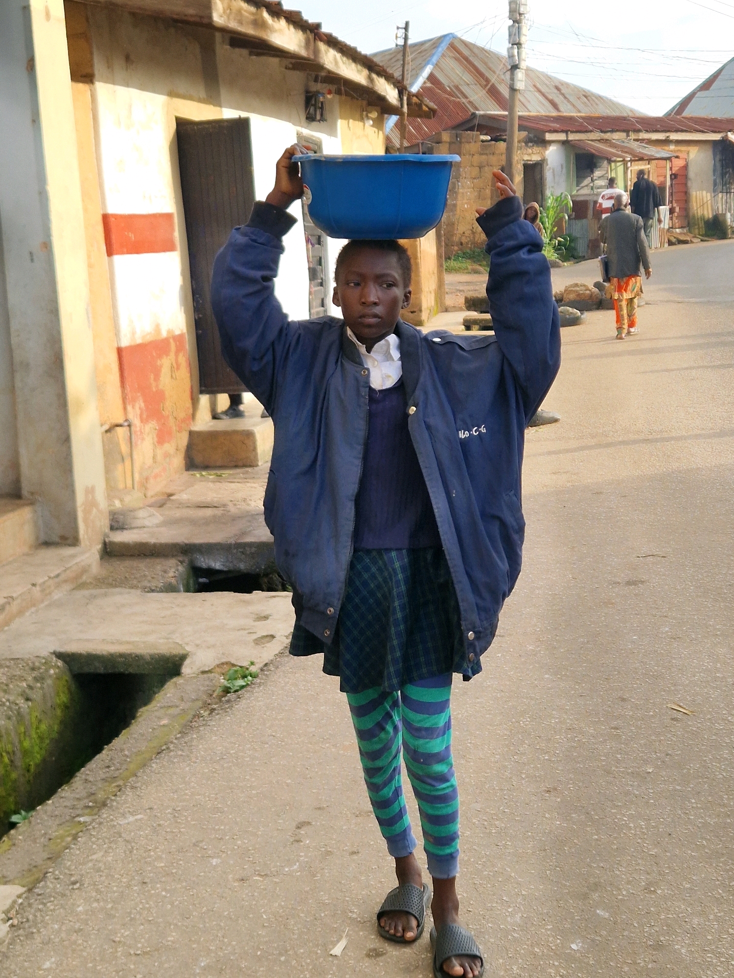 A young person, appearing to be of African descent and dressed in a blue jacket over a dark sweater and a green and blue striped skirt or culottes, is walking forward on a paved street, balancing a large blue basin on their head. They are wearing dark gray slide sandals. The person's face is visible, showing a neutral expression. In the background, there are buildings with corrugated metal roofs and some structures that appear to be made of brick and concrete. The environment suggests a street scene in a town or city, possibly in Nigeria. The buildings and street are somewhat weathered, with visible signs of use and age. To the left of the person, there is a concrete drainage ditch. Further down the street, two adults are walking away from the camera, one in a dark jacket and colorful patterned pants, and another in a dark jacket. The lighting indicates it is daytime, with sunlight casting shadows.