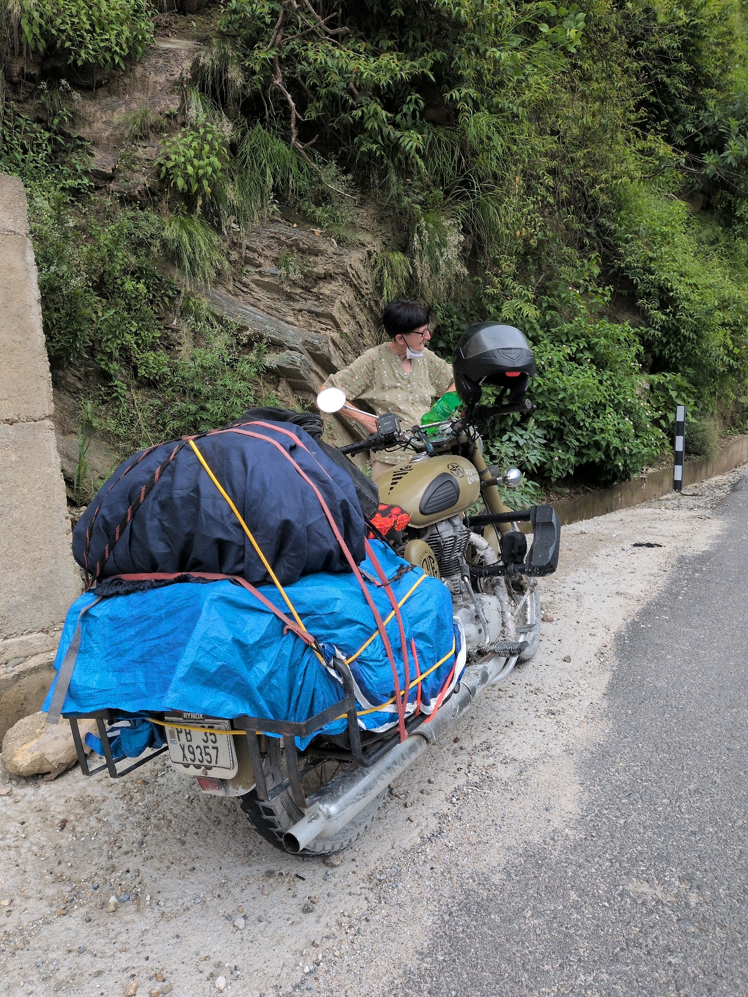 In Uba, India, a person wearing a light brown shirt and black glasses stands next to a loaded motorcycle. The motorcycle is olive green and has a black helmet resting on its handlebars. The rear of the motorcycle is covered with a blue tarp, secured with red and yellow straps, and a larger dark blue bag is strapped on top of the tarp. The license plate of the motorcycle reads "PB 35 X9357". The scene is located on a dirt and gravel shoulder next to an asphalt road, with a steep, vegetated hillside on the left.