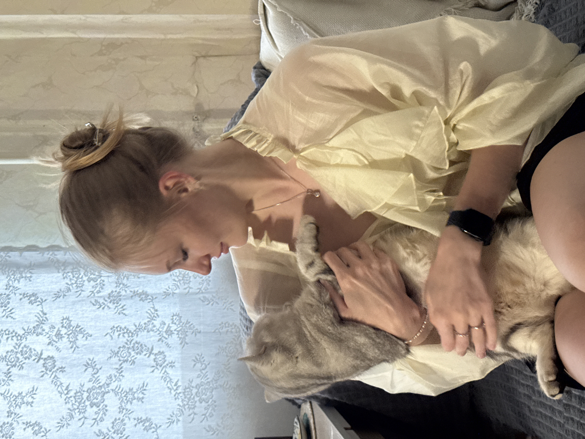 A woman is pictured holding a cat while sitting on a bed. The woman has her hair pulled up in a bun and is wearing a light-colored ruffled blouse and black shorts. She is wearing jewelry on her wrist and fingers, including a smartwatch. The cat is grey with tabby markings and appears to be sleeping. The background shows a patterned curtain and bedding. The scene is likely indoors, and the lighting suggests it could be daytime. The overall mood appears to be relaxed and affectionate. The location context is Perm, Russia. There is no visible text in the image.