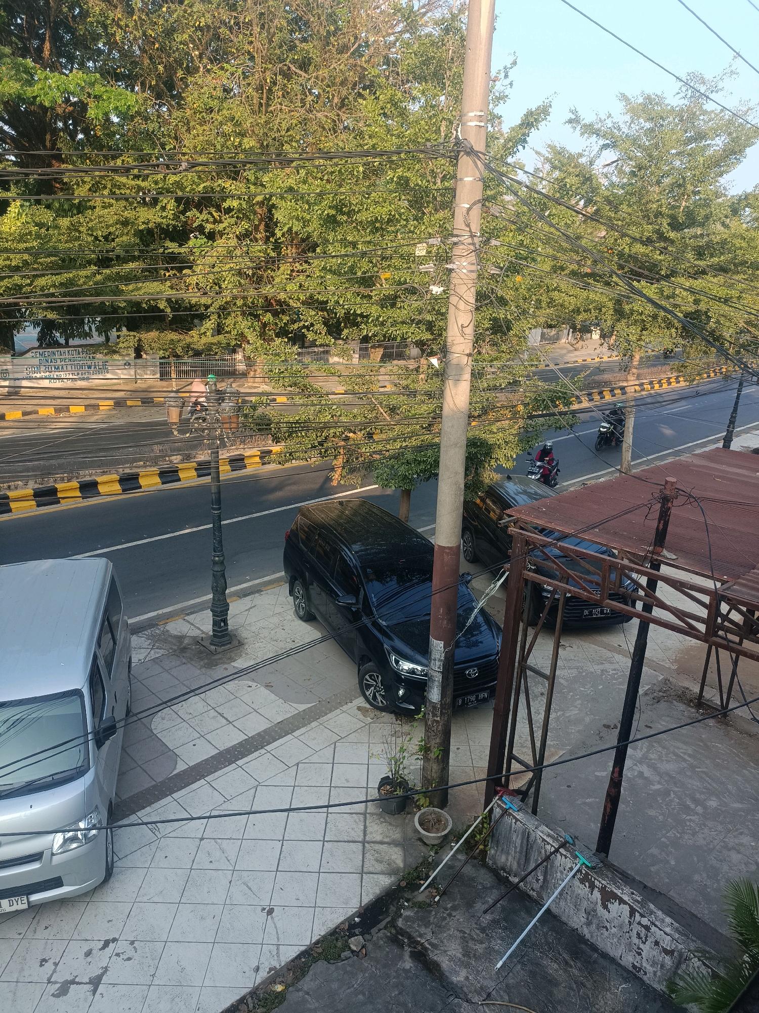The image depicts a street scene in Palopo, Indonesia, on a clear, sunny day. A black SUV and a silver van are parked on a tiled sidewalk adjacent to a road. The road is marked with yellow and black striped barriers and is traversed by two motorcycles. Power lines crisscross the frame, supported by a tall utility pole. Trees with lush green foliage line the street, and a lamppost stands on the sidewalk. In the background, a building with signage indicating it is part of the "PEMERINTAH KOTA PALOPO DINAS PENDIDIKAN" (Palopo City Government, Education Department) is visible. In the foreground, a dilapidated structure with a rusty, corrugated metal roof is present, along with cleaning tools leaning against a wall.