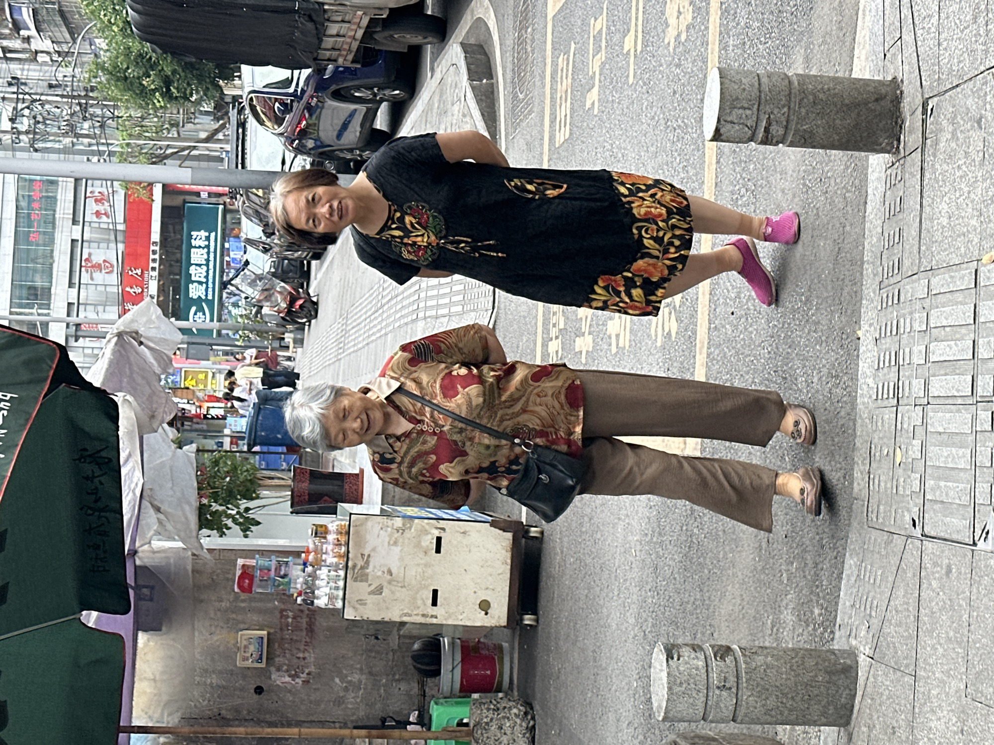 Two elderly women stand on a city street in Jiangjin, China. One woman wears a black dress with floral trim and pink shoes. The other woman wears a patterned blouse, brown pants, and carries a black purse. They are positioned near a food stall under a green umbrella and beside concrete posts in the street. Buildings are visible in the background.