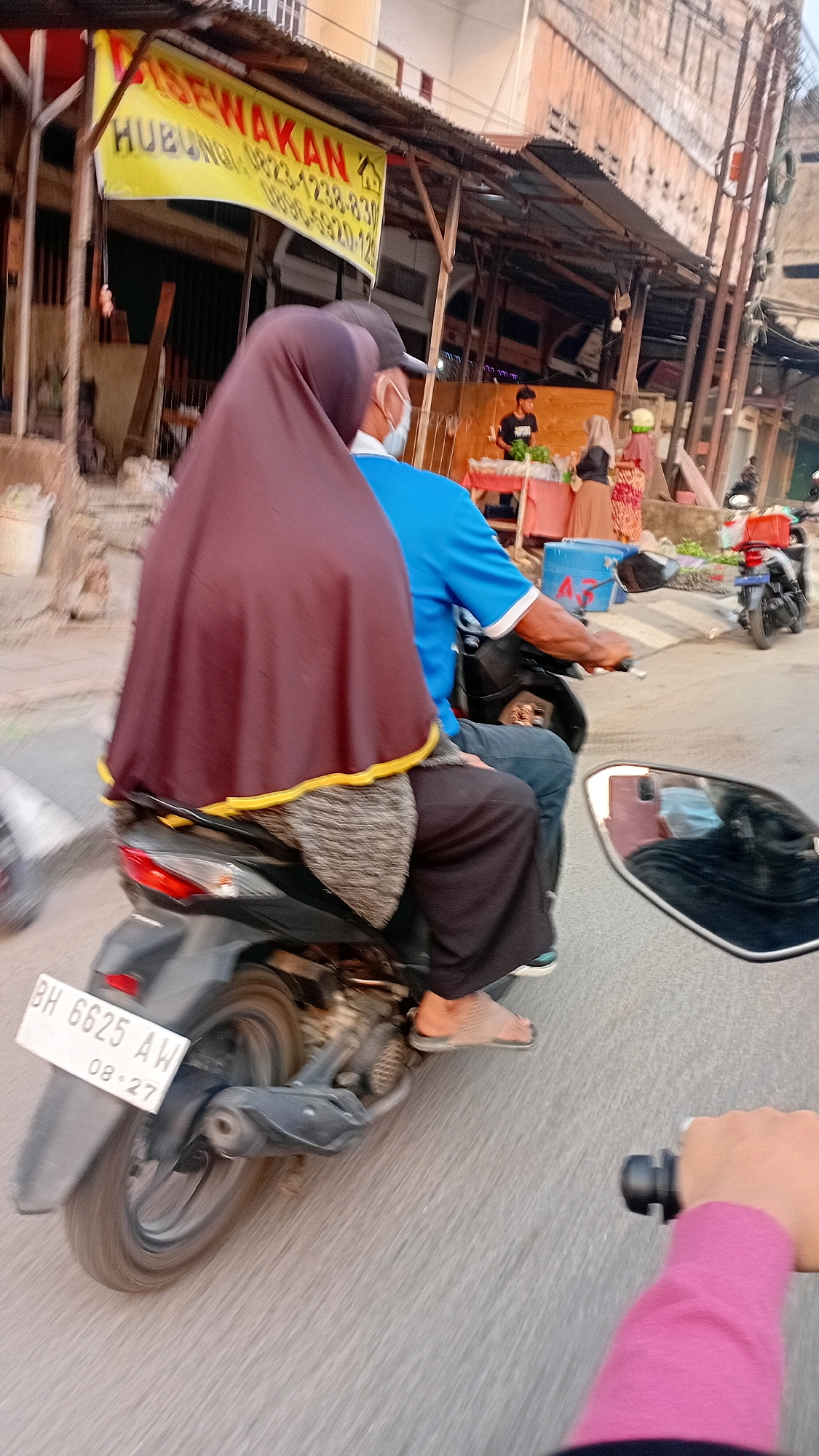 In Jambi, Indonesia, a motorcyclist wearing a blue shirt and black pants rides down a street with a passenger behind him wearing a dark purple hijab. The passenger's left arm is visible, with a grey and black patterned fabric draped over their back. The motorcycle's license plate reads "BH 6625 AW" and "08.27." In the background, a yellow banner displays "DISEWAKAN HUBUNGI 0823-238-830 0896-5920-128" along with a house icon. A market stall is visible with people selling vegetables. The environment appears to be a busy urban street with buildings and other motorcycles.