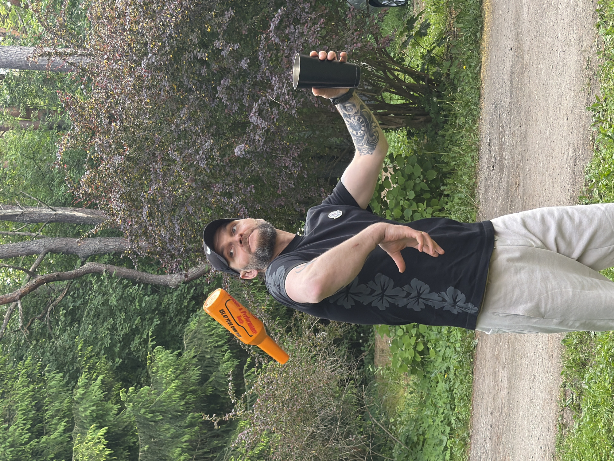 A man with a beard wearing a black t-shirt, a black baseball cap, and light-colored pants is standing in a park-like outdoor setting. He is looking up and to the left, with his right arm extended and holding a black cup. An orange object, resembling a bottle with text on it, is suspended in the air to his left. The background consists of green trees and bushes, with a dirt path in the foreground. The scene appears to be in a public park.