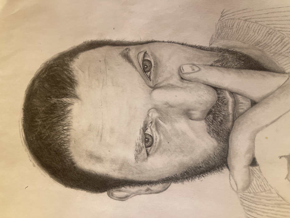 The image displays a detailed pencil drawing of a male individual, rendered in grayscale on a light-colored paper surface. The portrait captures the subject from the chest up, focusing on his head and shoulders. He has short, dark hair and a full beard, both depicted with visible texture. His eyes are open and directed forward, with a direct gaze; the eye on the viewer's right appears slightly more narrowed than the left. The man's left hand (viewer's right) is raised, with his index finger positioned vertically against the side of his nose, near the nostril and extending towards the upper lip. The thumb and other fingers are partially visible, curled beneath his chin. He is wearing a garment with horizontal ribbed or striped detailing, visible across his shoulders and upper chest. The background is a plain, unadorned surface, consistent with drawing paper, showing minor creases.
