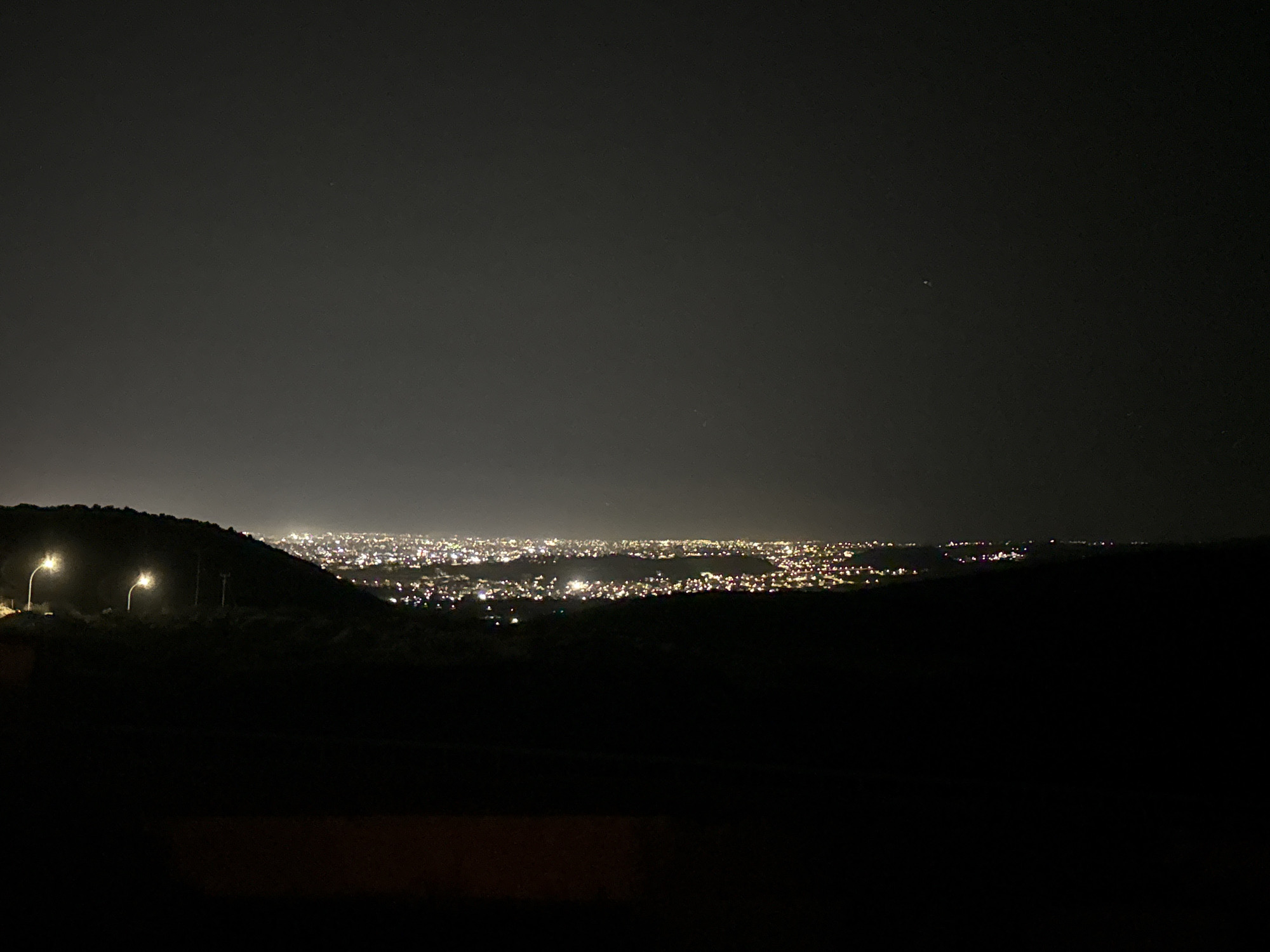A nighttime, wide-angle shot captures a panoramic view of a sprawling city illuminated by countless lights, stretching across the horizon. The city lights form a bright band against the dark landscape, suggesting a significant urban area. In the foreground, a dark, silhouetted hill rises, with two prominent streetlights casting pools of yellow light onto its slopes. The sky above is a deep, dark gray, with a faint scattering of stars visible, and a single, small, bright object is discernible in the upper right quadrant of the image. The overall scene is one of a peaceful, illuminated urban expanse viewed from a higher vantage point on a dark night. The image is likely taken from a vantage point overlooking the Agios Tychona Community in Cyprus, showcasing its nighttime glow.