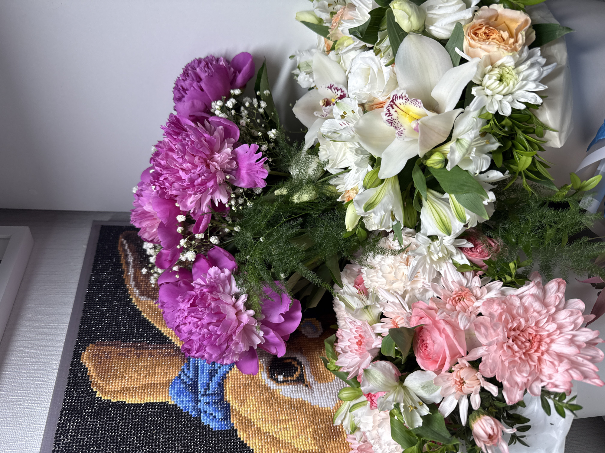 The image displays a detailed still-life composition featuring multiple bouquets of fresh cut flowers and a framed diamond painting.
In the foreground and midground, three distinct floral arrangements are prominently positioned. The upper bouquet consists primarily of vibrant fuchsia/magenta peonies, interspersed with small white Gypsophila (baby's breath) and fine green fern-like foliage. Below this, on the right, a second large bouquet showcases several white orchids with distinctive yellow and maroon speckled centers, accompanied by white Alstroemeria (Peruvian lilies), white roses, white chrysanthemums, and a single peach-colored garden rose. The third bouquet, located in the lower-left portion of the frame, features various shades of pink and white flowers, including light pink chrysanthemums, lighter pink carnations, pink spray roses, and white Alstroemeria. All bouquets are wrapped at their bases with white or light-colored material, partially obscuring their lower stems.
In the background, to the left, a framed diamond painting is visible. The artwork depicts the head and upper body of a light brown or tan animal, consistent with a rabbit, wearing a bright blue bow tie or scarf. The rabbit has a visible eye with a dark pupil and white highlights. The background of the diamond painting is solid black, composed of numerous small, uniformly dark diamond drills, whose individual facets are discernible. The painting is encased in a light-colored frame. Above the diamond painting, a section of a textured, light-colored wall or wallpaper is visible, leading to a white horizontal trim element. The wall behind the floral arrangements appears plain and light grey or white. No individuals are present in the scene, and no actions are depicted. The lighting is uniform and bright throughout the composition.
