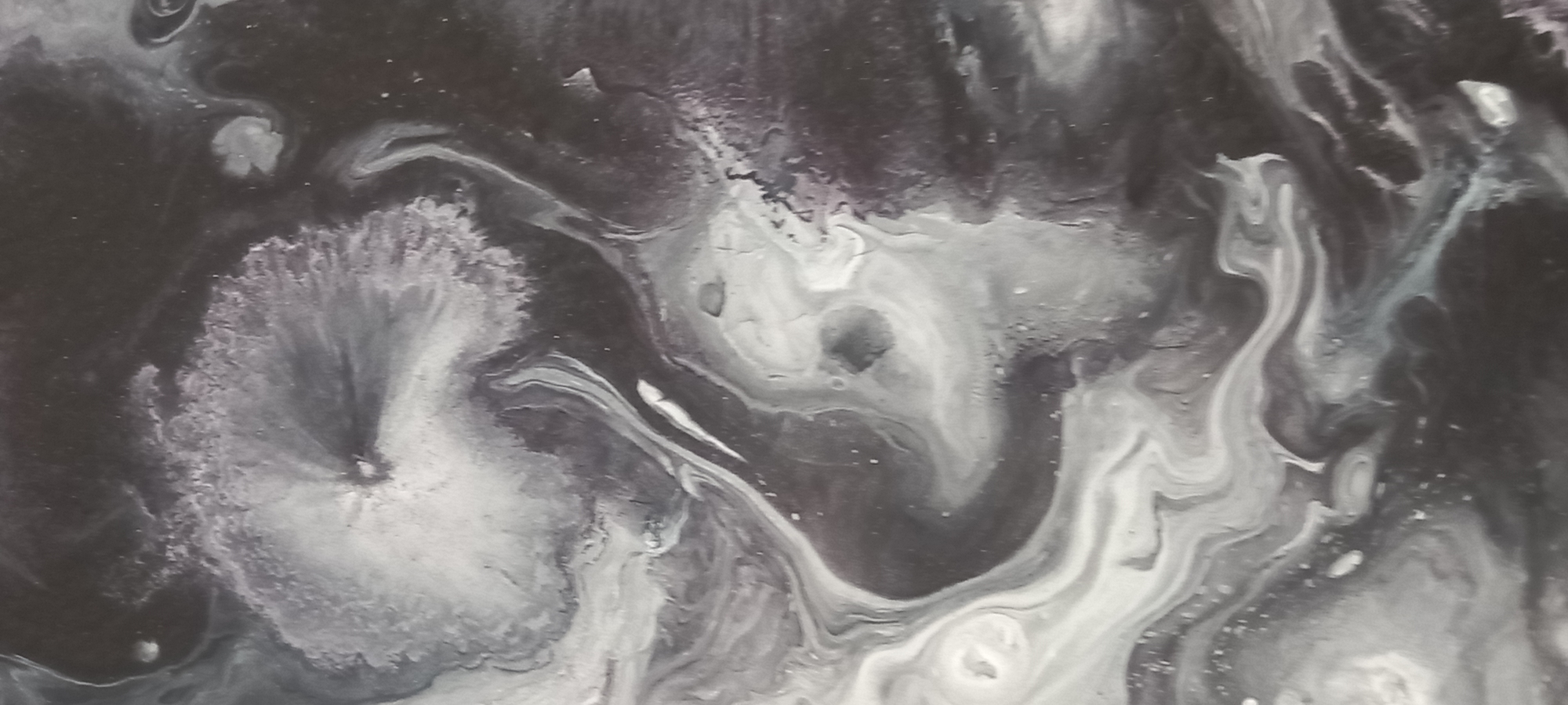This is a close-up, abstract photograph of what appears to be a fluid art painting. The image is dominated by swirling patterns of black and white paint, creating a marbled or nebula-like effect. There are areas of dense black, interspersed with streaks and clouds of white and lighter gray. Some areas show a textured, almost cracked appearance, while others are smooth and fluid. The lighting is soft, casting subtle shadows and highlighting the glossy sheen of the paint in places. The photograph does not contain any recognizable landmarks or elements that would allow for identification of a specific location within Kryvyi Rih, Ukraine. There are no people, animals, or identifiable objects present. The focus is solely on the abstract patterns and textures of the artwork.