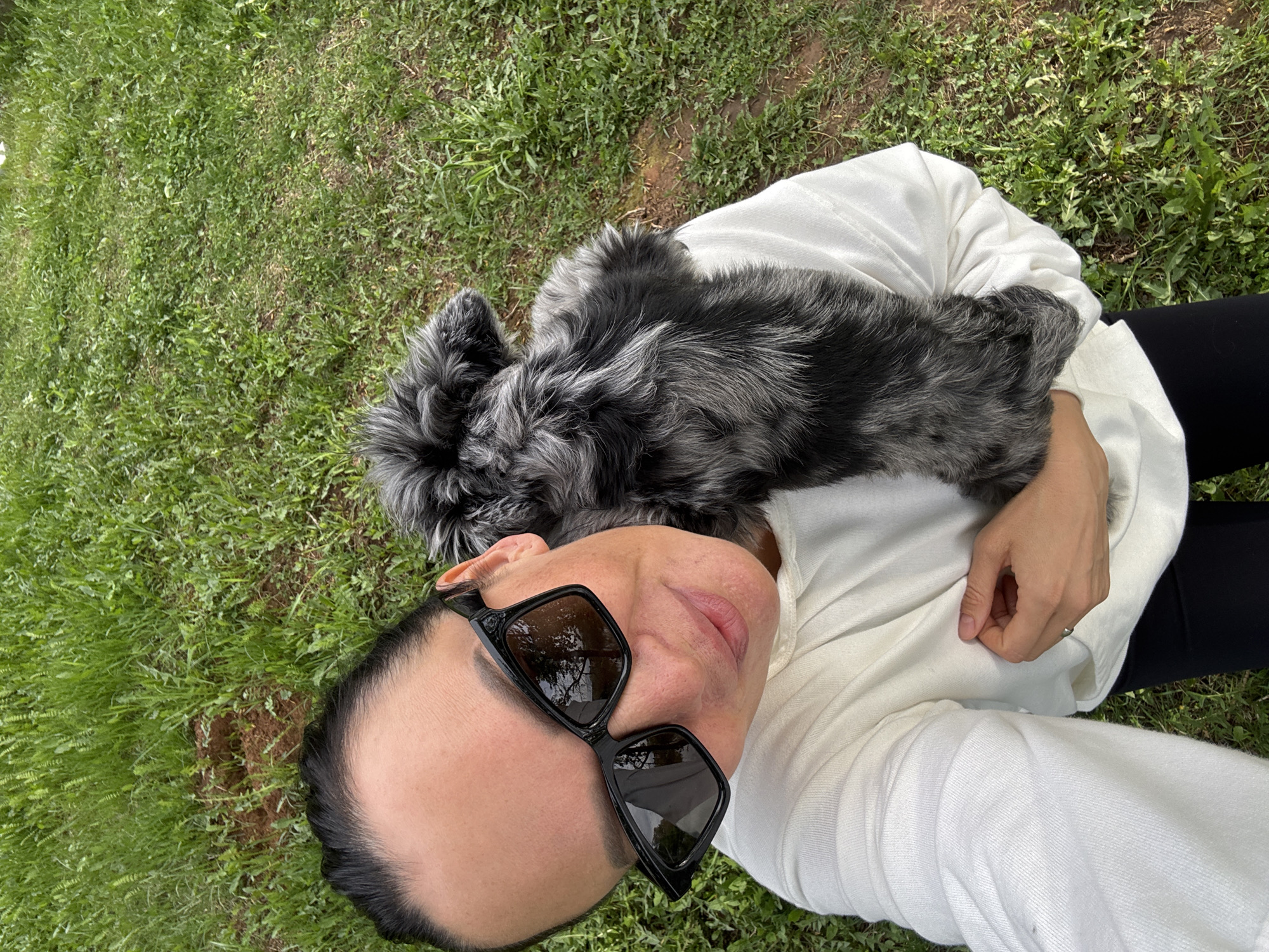 A woman lies on green grass, holding a gray and black dog. She wears black pants, a white shirt and black sunglasses. The location is not identifiable as Chaikovsky, Russia from the visual data.
