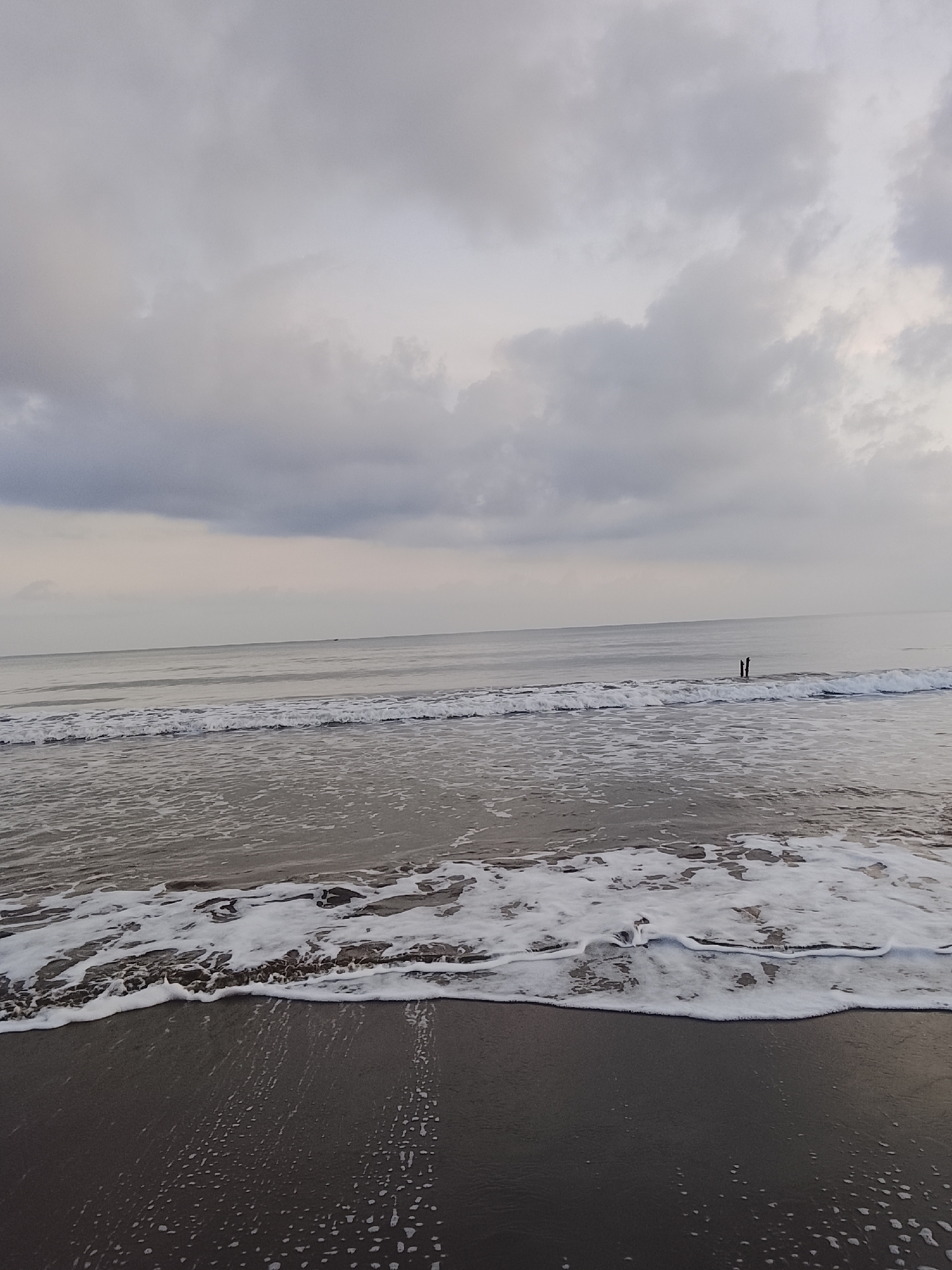 The image depicts a tranquil beach scene at dawn or dusk in Reudeuep, Indonesia. The sky is overcast with soft, diffused light filtering through thick gray clouds. Gentle waves with frothy white foam are rolling onto a dark, wet sand beach. In the middle ground of the ocean, two figures are standing in the water, appearing as small, dark silhouettes against the lighter horizon. The overall atmosphere is serene and still.