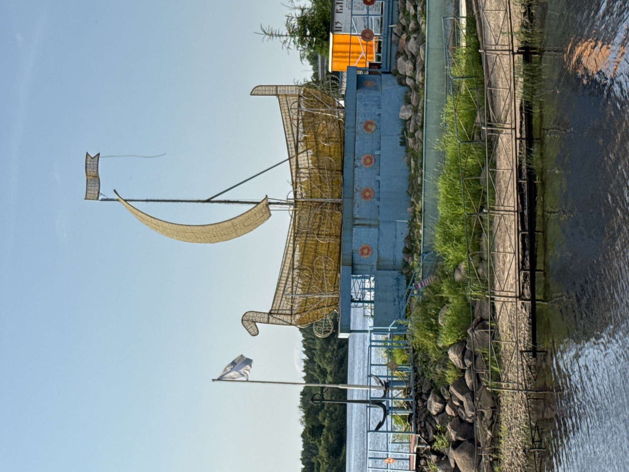 In Kirishi, Russia, a decorative Viking ship sculpture stands on a blue platform by the Volkhov River. The sculpture is made of yellow metal mesh and features a mast with a flag. A large anchor is positioned near the sculpture on the left side. The scene is set against a clear blue sky and a backdrop of lush green trees. A small wooden bridge extends over the water in the foreground. Orange life preservers are visible on the blue platform.