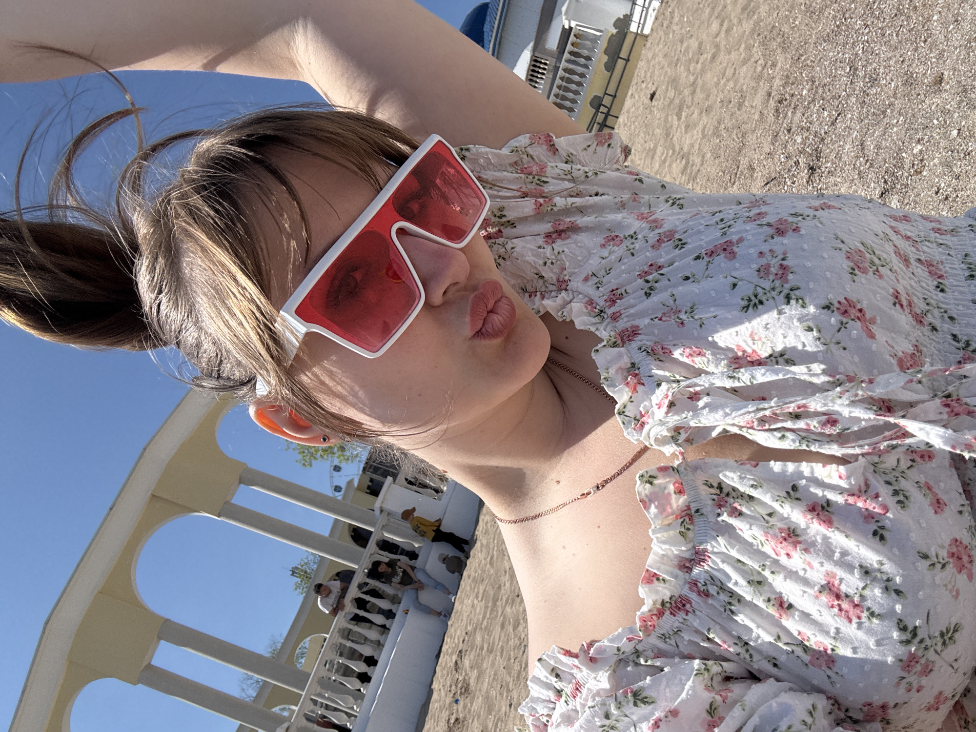 A woman poses for a selfie, outdoors on a beach in Yevpatoriya, Ukraine. She is wearing red-tinted sunglasses with white frames and a white floral-print dress. Her hair is pulled back in a ponytail. In the background, there is a white colonnade structure with a beach and people visible behind the colonnade. The sky is clear and blue.