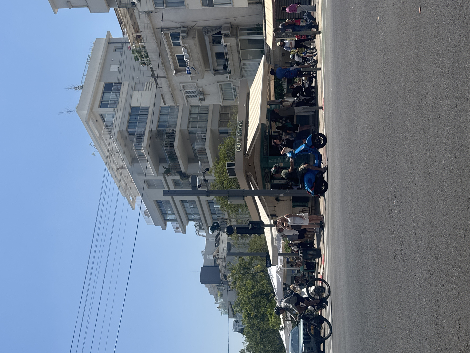 The image depicts a street scene in Tel Aviv, Israel. A multi-story white building with balconies lines the left side of the frame, adjacent to a sidewalk cafe named "Cafe Kiosk". Patrons sit at tables under an awning. Several people stand on the sidewalk and several blue scooters are seen. Bicycles are parked along the street curb. Power lines stretch across the upper left corner of the frame against a clear blue sky. The street itself, occupying a significant portion of the right side of the image, is paved in asphalt.