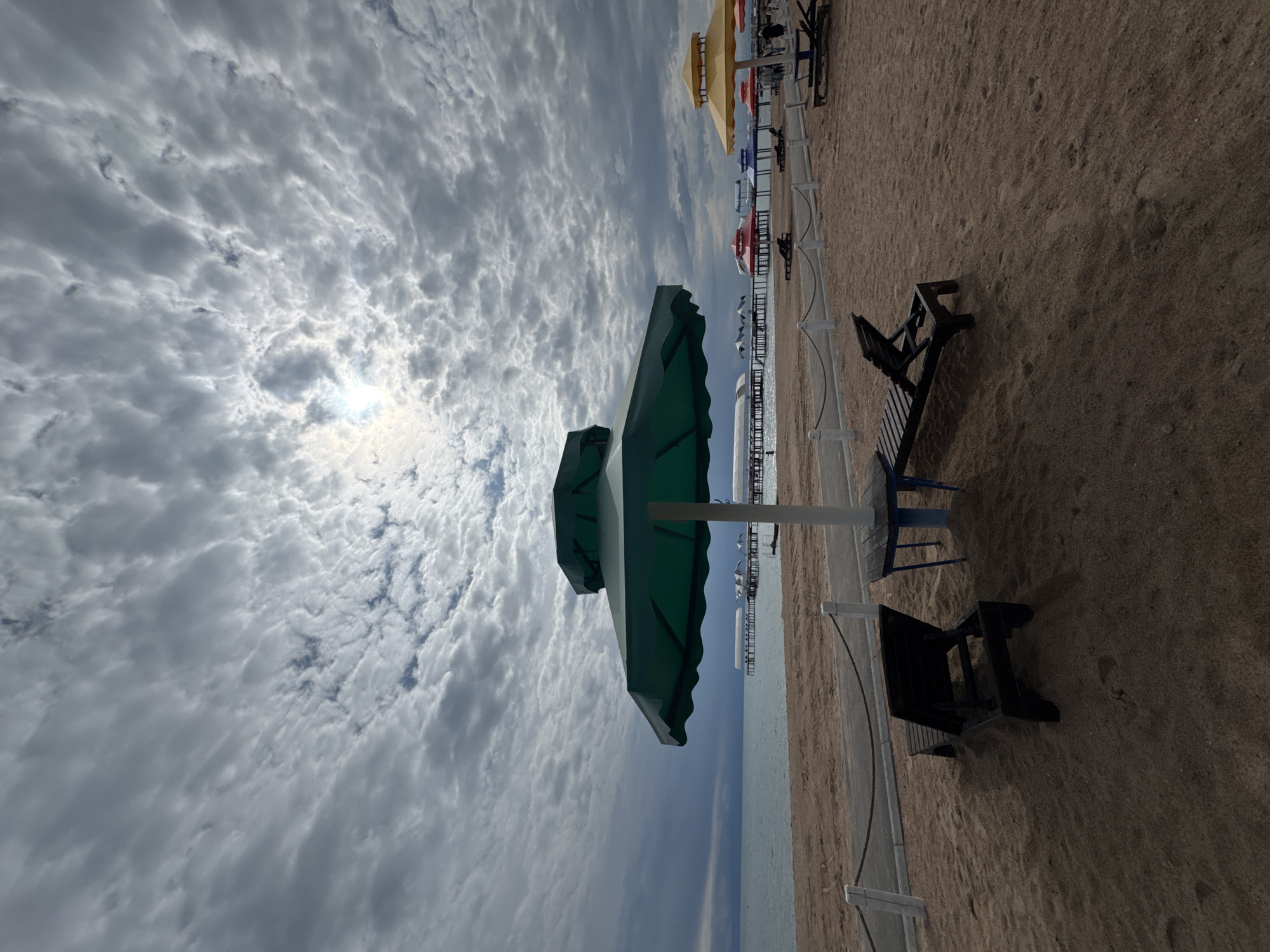The image depicts a beach scene in Sary-Oy, Kyrgyzstan. In the foreground, a dark wooden lounge chair is positioned on the sandy beach, angled slightly towards the viewer. Another identical lounge chair is partially visible to its right, also on the sand. A large, dark green beach umbrella dominates the center of the frame, casting a shadow. A small blue table with a blue pole stands beneath the umbrella. Further into the background, a paved path runs parallel to the beach, separated by a low white fence with a rope railing. Beyond the path, several colorful beach umbrellas, including red, blue, and yellow ones, are set up, with lounge chairs beneath some of them. People are visible in the distance, walking near the umbrellas. A series of piers or jetties extend into the calm, light blue water, with structures on them. The sky is overcast with dramatic, textured clouds, and the sun is partially visible through a break in the clouds, creating a diffused light. The overall impression is a quiet, somewhat cloudy day at a resort beach.